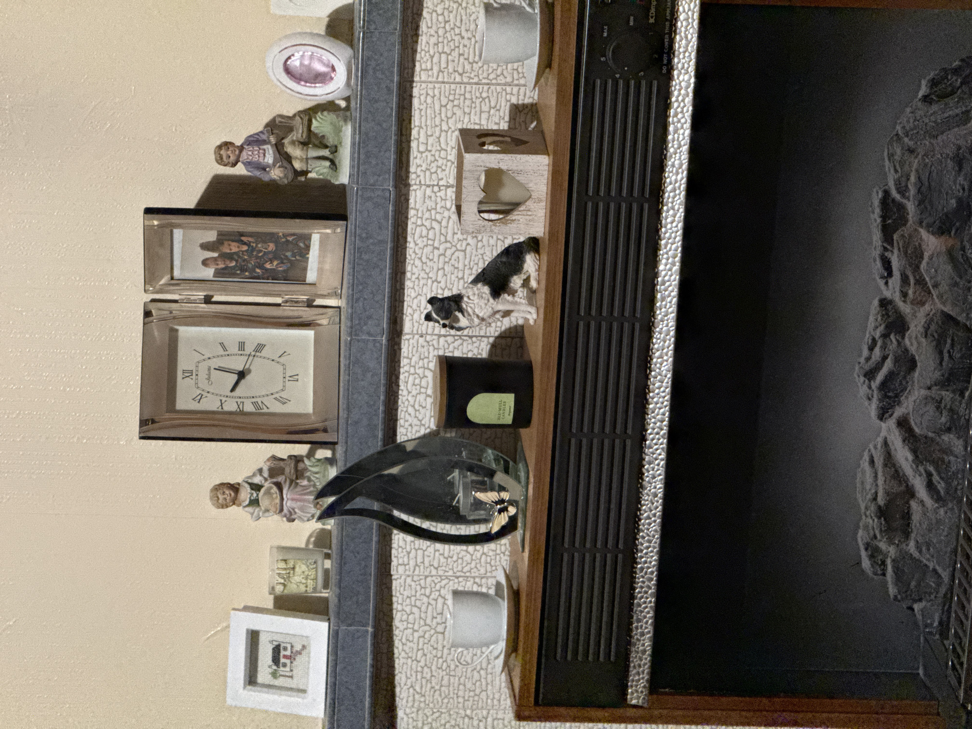 A still life image reveals a diverse collection of items arranged on a shelf. At the top, a small, ceramic figurine of a child with a sheep sits next to a decorative dish. Below, a framed picture and a silver-trimmed clock with Roman numerals face the viewer. Further down, a porcelain figurine of a woman with a basket is accompanied by a mug and a small, framed picture.
Adjacent to the shelf's contents is a small, white tissue box with heart-shaped cutouts, and a figurine of a black and white dog. There is also a black jar candle, a decorative glass butterfly on a stand, and a mug. To the right, an electric fireplace unit is visible. The unit has a black grille-covered section and a simulation of a stone wall. The unit's control panel is visible on the top of the grille.