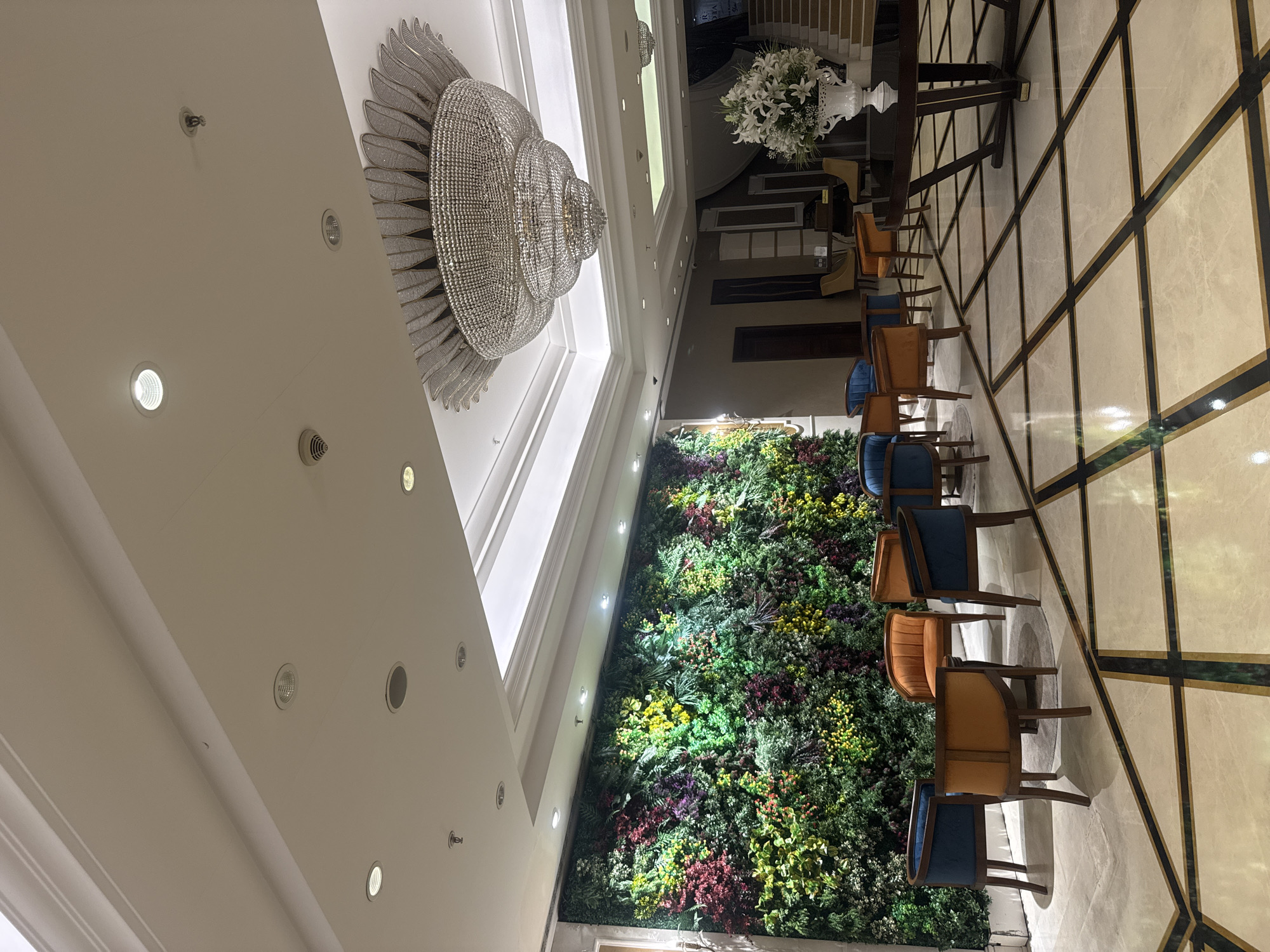 The image shows an interior space in what appears to be a hotel or large building in Bağcılar, Turkey. A large, ornate chandelier hangs from the ceiling. Along one wall is a vertical garden or wall of greenery. There are a row of chairs upholstered in alternating blue and orange fabric, arranged near the green wall. The floor is tiled in a pattern of light colored stone with dark accents forming a grid. The overall impression is of a modern, well-lit lobby or waiting area.
