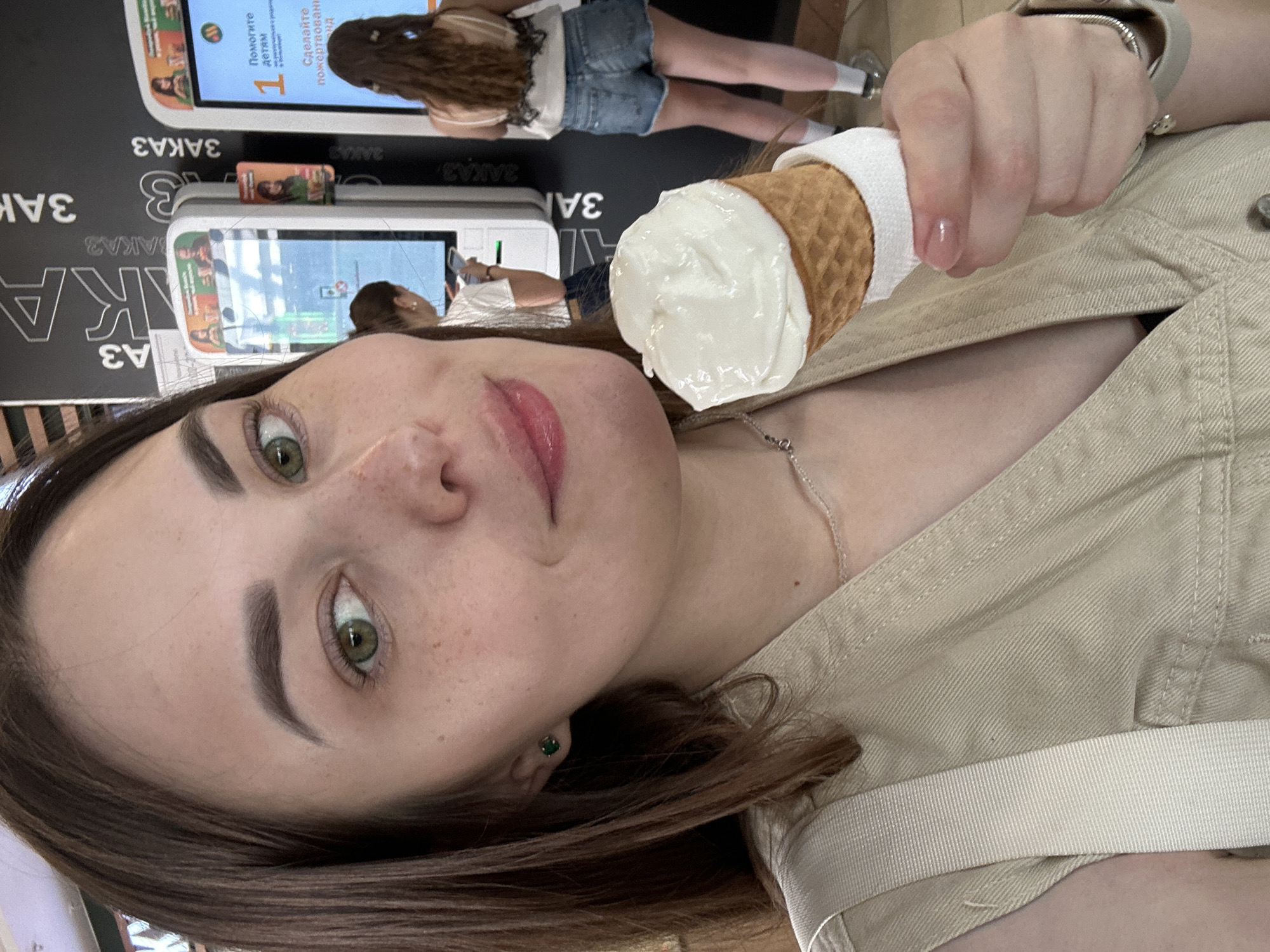 A selfie shows a young woman with green eyes and brown hair, facing the camera. She is holding an ice cream cone with a white-colored ice cream scoop. The cone is wrapped with a white napkin. She is wearing a light-colored top and a thin silver necklace. Her lips are glossy pink. Behind her are self-service kiosks with digital displays. Other people are seen in the background. The signs on the kiosks are in Russian. The location is in Perm, Russia.