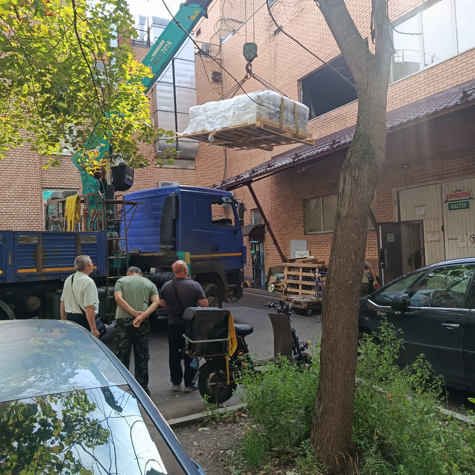 A crane is lifting a pallet of materials wrapped in plastic. A blue truck with a crane is parked on the street. Three men are standing near the truck, seemingly supervising the operation. One man in a light colored shirt and black bag across his chest is looking towards the crane. Another man in a green shirt and camouflage pants has his hands behind his back and is looking down. A third man in a dark shirt is standing with his back mostly to the camera. A dark colored car is parked on the right side of the image, and a black motorcycle with a yellow bag is parked behind the truck. The scene appears to be an outdoor industrial or commercial area with a brick building in the background. The sun is shining, casting shadows, suggesting it's daytime. A sign on the building reads "ДОМАШНИЙ МАСТЕР" (DOMASHNIY MASTER) which translates to "HOME MASTER." The weather appears to be pleasant.