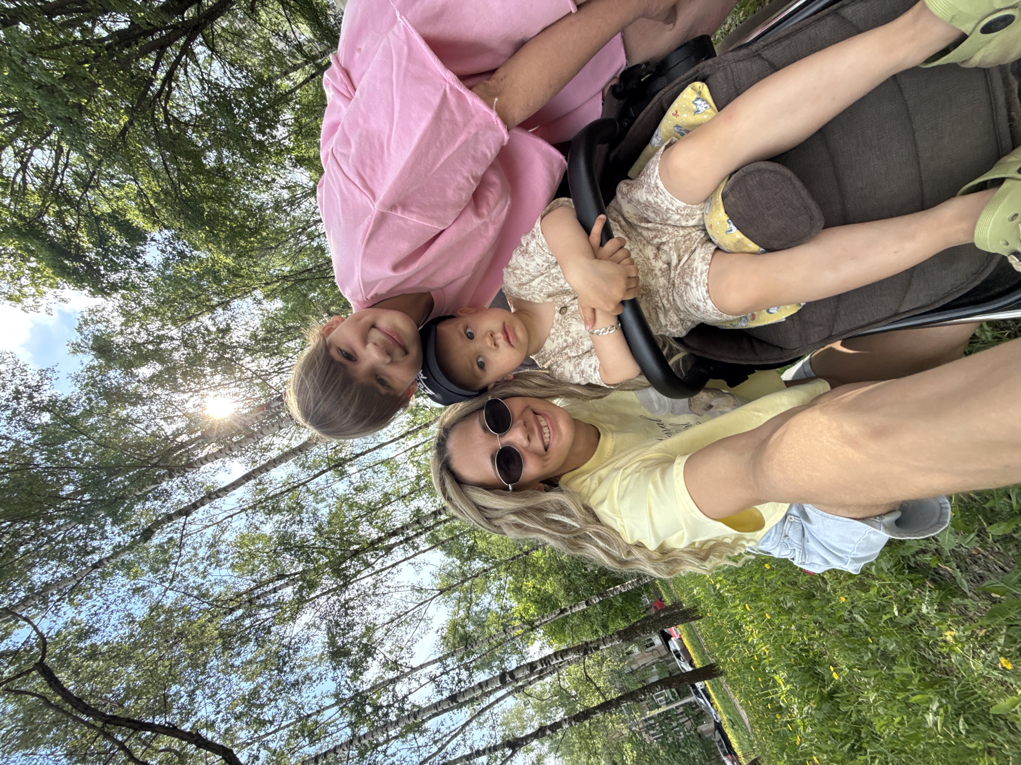 Four people – three children and an adult – take a selfie outdoors. The baby is in a stroller. They are surrounded by a green, sunlit park with birch trees. In the background, there are buildings, roads, and cars that indicate the location is in the city. It may be Rusanovo, Russia.