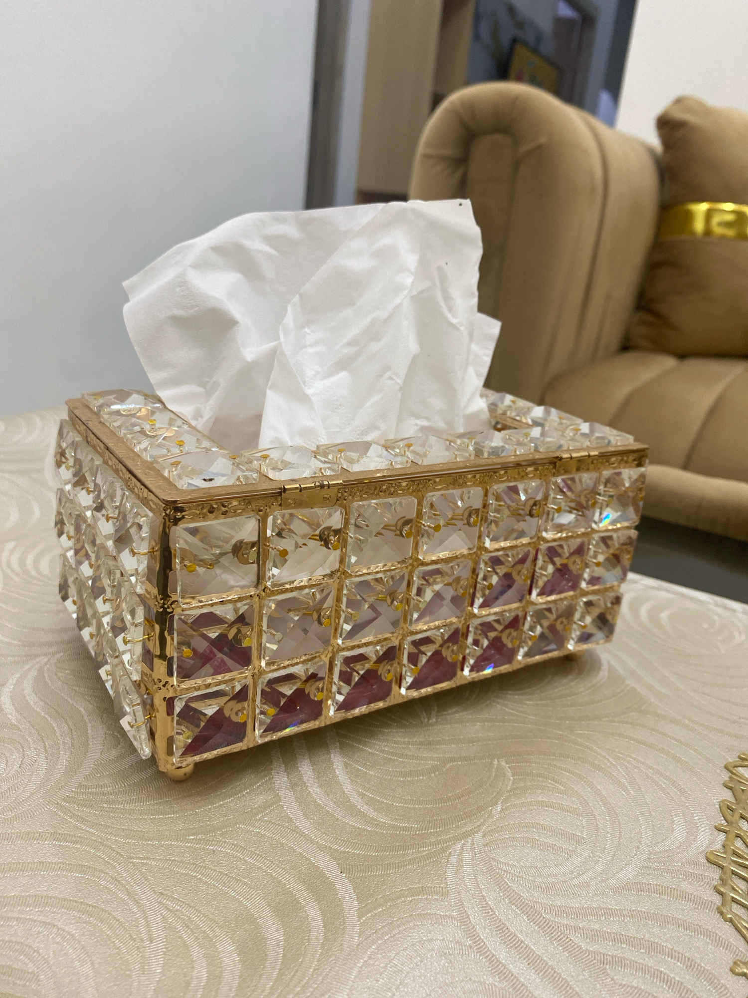 A close-up, eye-level, indoor shot displays a decorative tissue box filled with white tissues. The tissue box is rectangular with a gold-colored metal frame and is encrusted with numerous clear, faceted crystals. The crystals are arranged in a grid pattern on the sides of the box, with some reflecting a reddish-purple hue from within. The top of the box has a gold frame around the opening for the tissues. The tissue box rests on a light-colored tablecloth with an embossed swirling pattern. In the background, out of focus, is a plush beige sofa with a gold band detail on a pillow, and a glimpse of a room interior with soft lighting. The scene is well-lit, highlighting the sparkle of the crystals. The image was captured from a static perspective, with no discernable movement. There are no people or animals present in the image. The location is not identifiable as Kota Dalam, Indonesia, based on visual cues alone.