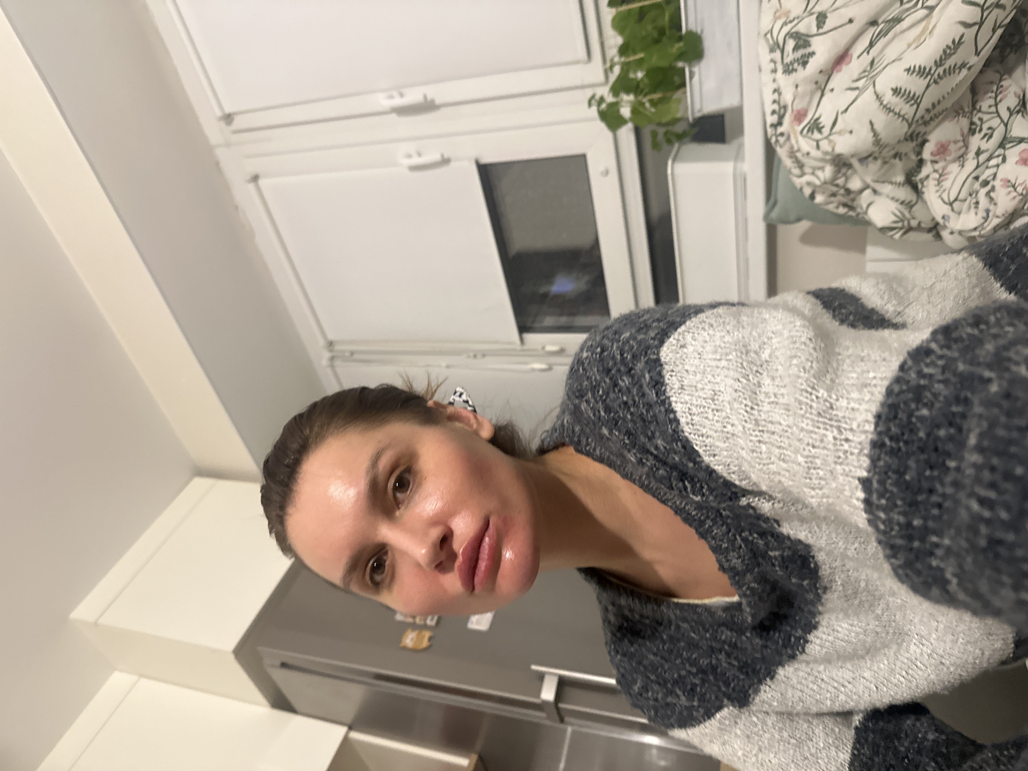 This close-up, portrait-style image captures a woman indoors, presented with the context that it was taken in Moscow, Russia. The woman, who appears to be in her 30s or 40s, is looking directly at the camera with a neutral expression. Her brown hair is pulled back, and her skin exhibits a slight sheen, particularly on her forehead and lips, which appear full. She is wearing a thick, textured cardigan or sweater with broad stripes or patterns in dark grey/blue and white tones.
The background suggests a modern home interior. To the woman's left and above her, white kitchen cabinets or ceiling panels are visible, along with the top portion of a silver-toned refrigerator. To her right, a white-framed window shows the dark exterior, indicating it is likely evening or night. A small, leafy green potted plant hangs or sits near the window. Further to the right, a bed or sofa is partially visible, covered with a white blanket adorned with a delicate floral pattern featuring green foliage and small pink flowers. The overall lighting is artificial, consistent with an indoor setting during darker hours. There is no discernible text visible in the image.
