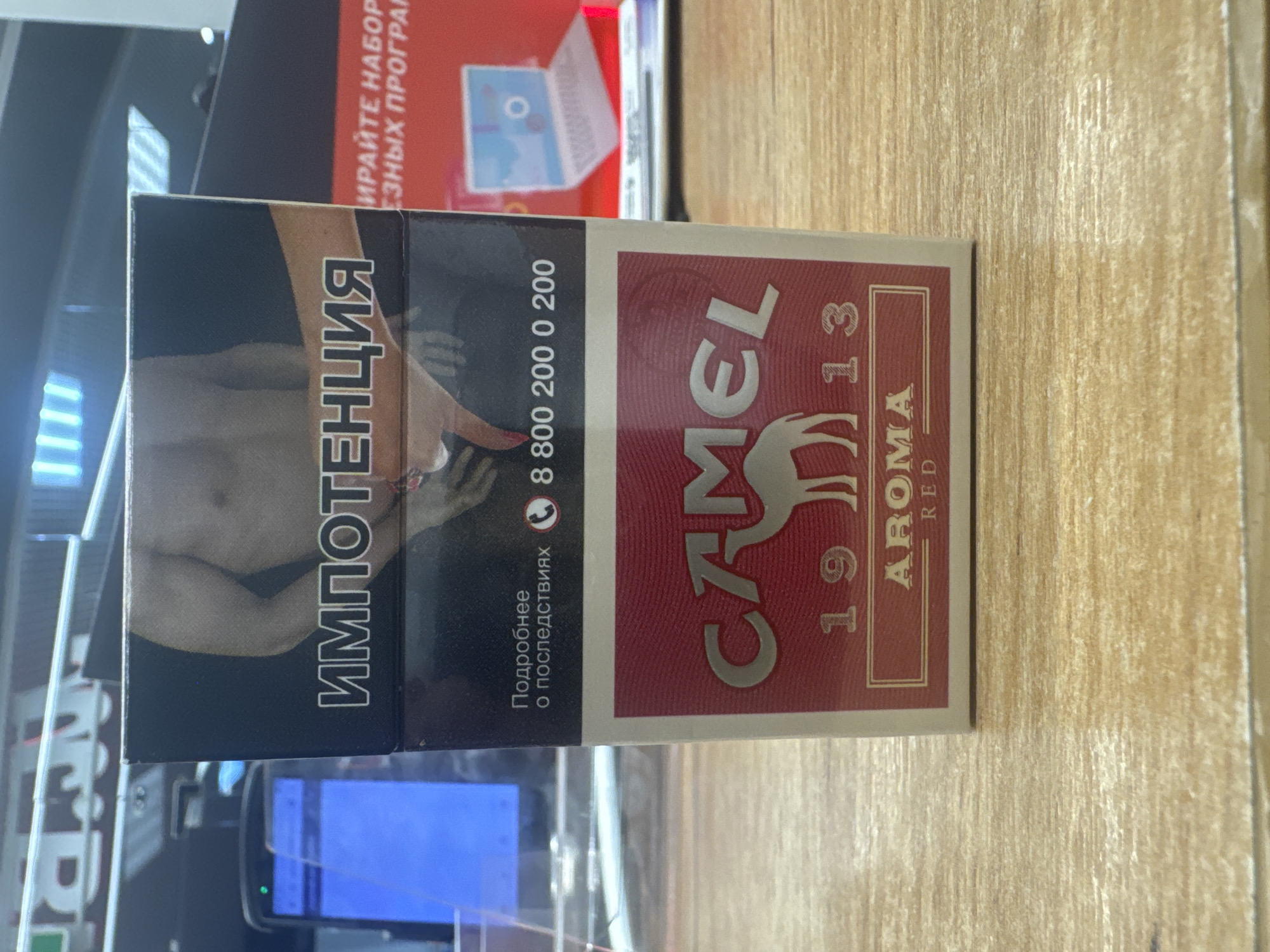 A close-up, eye-level, vertical shot showcases a Camel Red Aroma cigarette pack positioned on a wooden surface. The pack features a dark band across the top with the word "ИМПОТЕНЦИЯ" in white lettering and an image of a bare-chested man. Below this, "Подробнее о последствиях" (More about the consequences) is printed, accompanied by a phone icon and the number 8 800 200 0 200. The bottom portion of the pack is red with the Camel logo, including a camel silhouette and "19 13". Text at the very bottom reads "AROMA RED." In the background, a smartphone with a blue screen and a laptop on a red surface are visible. The scene is devoid of any discernible landmarks to determine its location within Ishim, Russia.