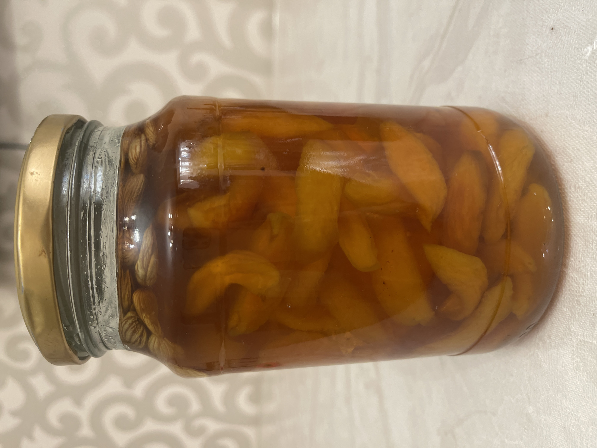 A medium shot displays a glass jar filled with preserved apricots and almonds in syrup. The jar has a golden lid and is placed on a white tablecloth with a subtle patterned background. The apricots are cut into slices and are a vibrant orange color, suspended in a dark amber syrup. Several almonds are visible at the top of the jar, submerged in the liquid. The lighting is soft, highlighting the textures and colors of the preserved fruit and the jar. The background features a light-colored, textured wallpaper with an abstract swirling pattern. No people are present in the image. The scene is static and appears to be a still life arrangement. This image was taken in Bishkek City, Kyrgyzstan.