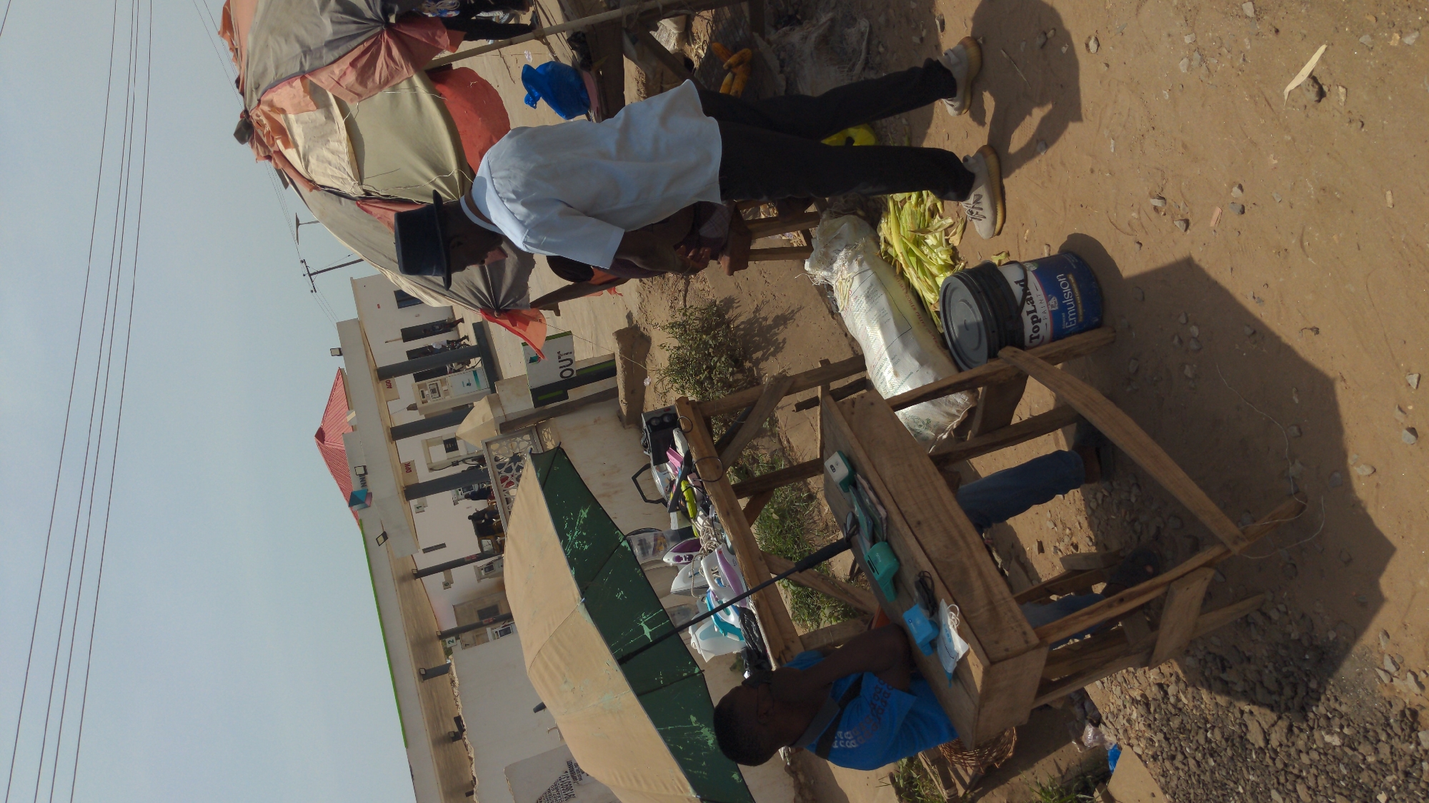 This image captures a vibrant outdoor scene, likely a market or roadside commercial area in Ado, Nigeria, under a bright, clear sky. The ground is a light brown dirt with scattered small stones, and strong shadows indicate sunny conditions, possibly around midday or early afternoon.
Several individuals are present, engaged in daily activities. A person wearing a white long-sleeved shirt, dark trousers, and a dark cap stands near a makeshift stall, appearing to attend to goods. To their left, another person in a blue t-shirt is seated behind a wooden display stand, looking downwards. Part of a third person's legs, clad in blue jeans, are visible beneath another wooden structure.
The scene is dominated by several vendor stalls. Two large umbrellas provide shade: one a mix of light-colored fabrics (beige, reddish), and another a prominent green. Crude wooden tables and stands serve as display units for various items. On one stand, an assortment of household goods such as multiple electric irons (in pink, white, and blue), kettles, and other small appliances are laid out. Nearby, a large white sack (possibly containing grain) and what appears to be corn on the cob or similar produce are visible. A blue bucket labeled "TopLand Paint" and "Emulsion" is also present on the ground next to a wooden stand.
In the background, a white commercial building with a red roof is visible. It features several structures that resemble fuel dispensers or pumps, suggesting it might be a petrol station. Against the light sky, multiple parallel power lines stretch across the frame. Visible text includes "OUT" on a sign and "TopLand Paint," "Emulsion" on the blue bucket.