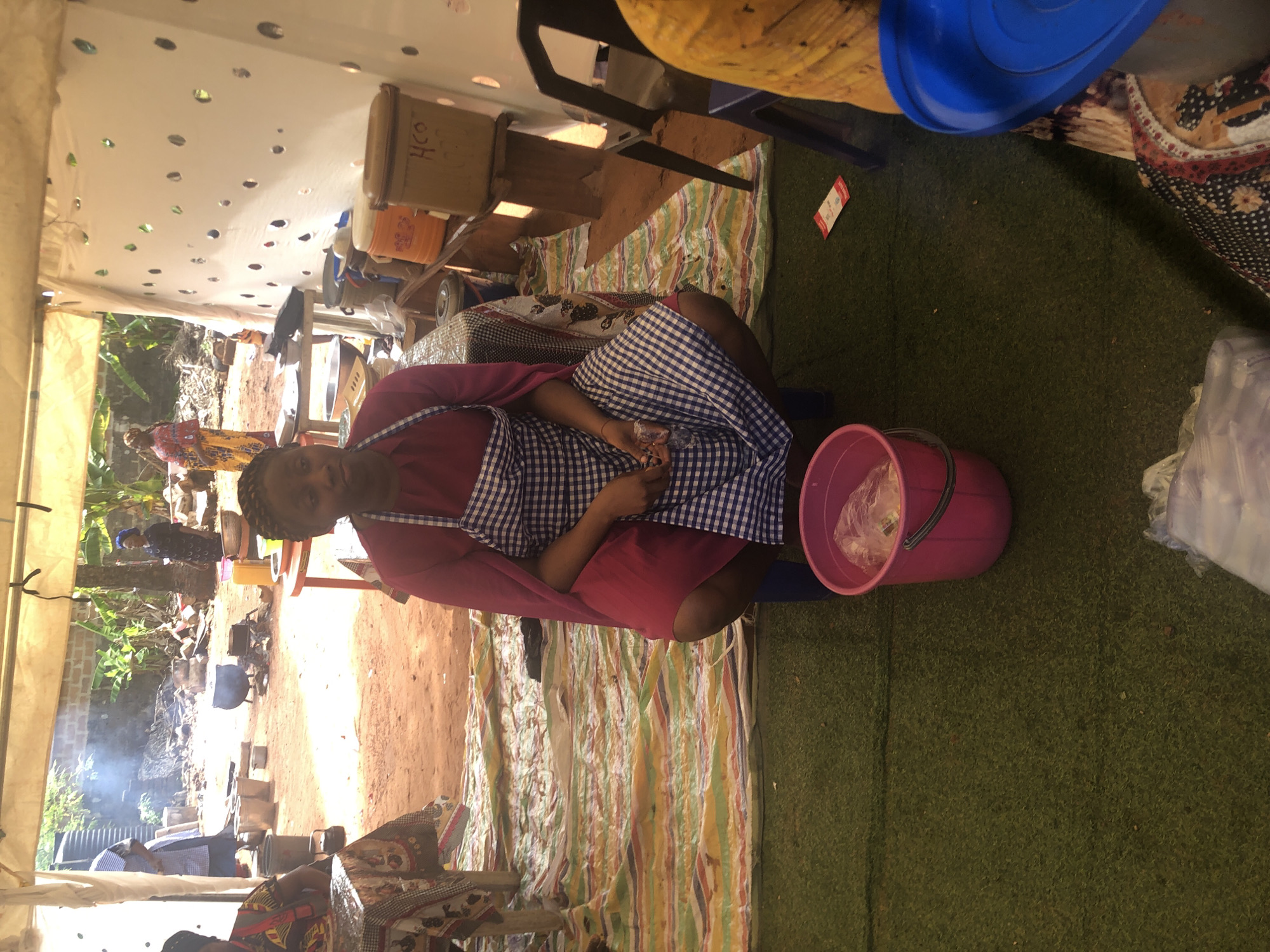 In Nteje, Nigeria, a woman wearing a red top and a blue and white checkered apron is seated under a tent. She is holding something in her hands, and a pink bucket is beside her. Outside the tent, there's a cooking area with pots on open fires. The tent's roof has numerous small holes, and the floor is covered with a striped rug and artificial turf.