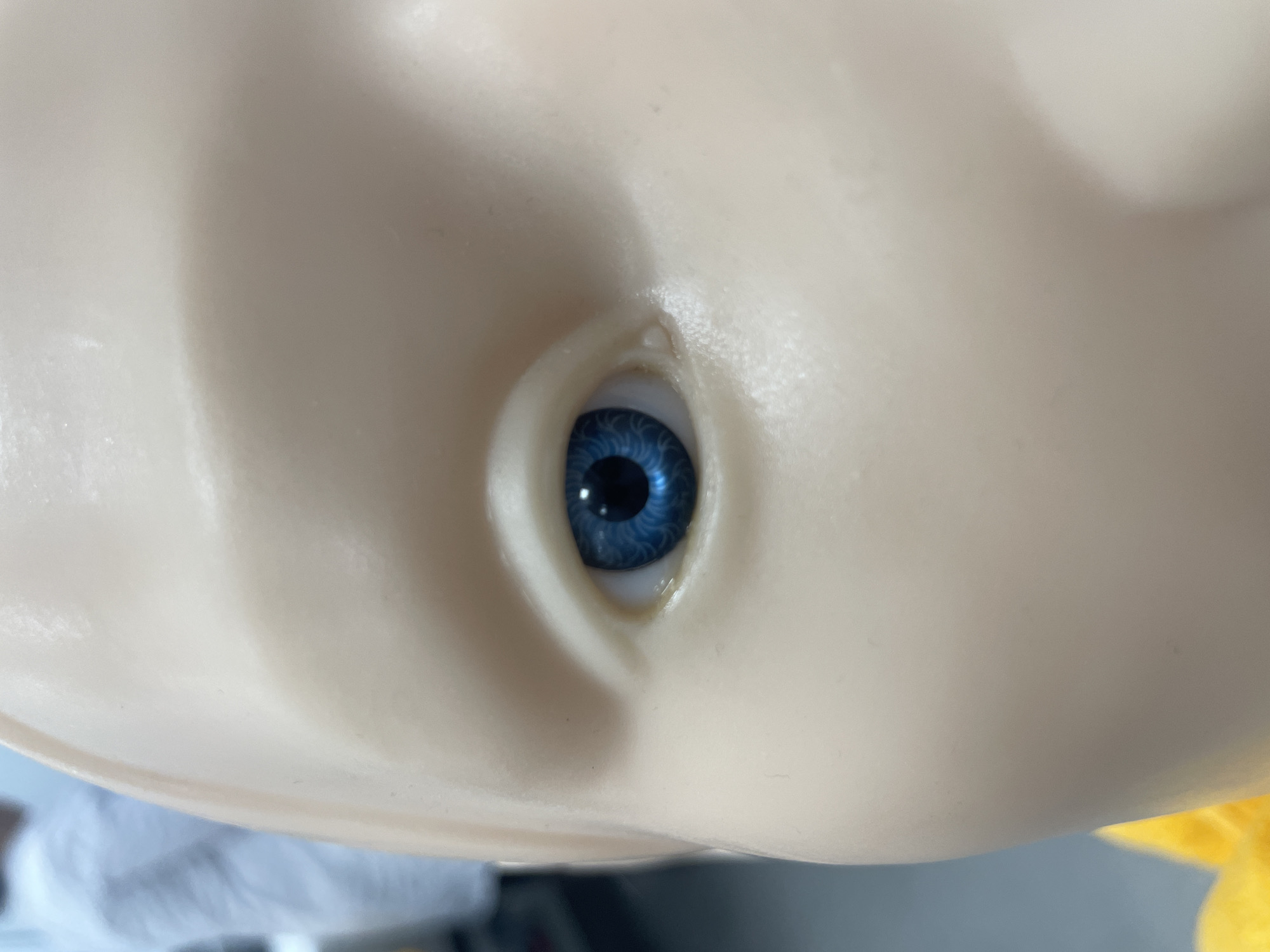 A close-up view of a doll's head, focusing on the left eye. The doll has a pale, fleshy complexion. The eye is bright blue, with a visible pupil and intricate iris details. The eye is set within a rounded eye socket. The overall impression is that the doll is plastic or made of a similar material.