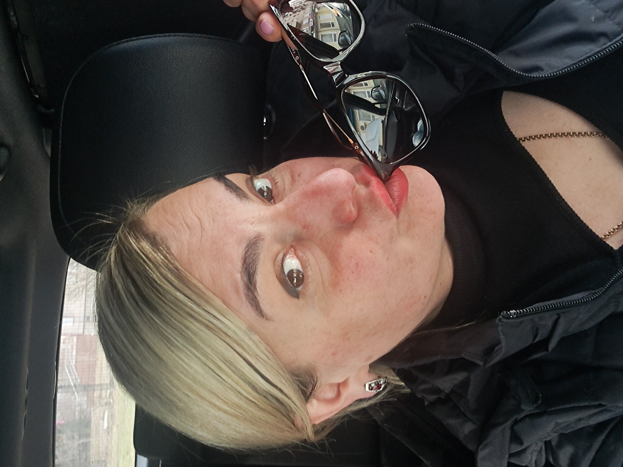 The image shows a blonde-haired Caucasian female, estimated to be an adult, situated in the passenger compartment of a vehicle in Elektrostal, Russia. Her left eye is open and directed at the viewer. Her lips are pursed, coated with red lipstick, and contact the edge of a pair of dark-lensed sunglasses held by her left hand. Her left hand displays light pink nail polish. She wears a dark, possibly black, quilted jacket over a dark top, with a visible gold-colored chain necklace. A silver-toned earring with a red stone is visible in her left ear. The vehicle interior is dark, with black upholstery visible on a headrest and the upper interior. Through the vehicle's rear window, a blurred exterior scene shows bare trees, green ground, and a distant light-colored building featuring a dark roof and external stairs. Reflections in the sunglasses depict architectural elements of a building exterior and a partial interior view of a car.