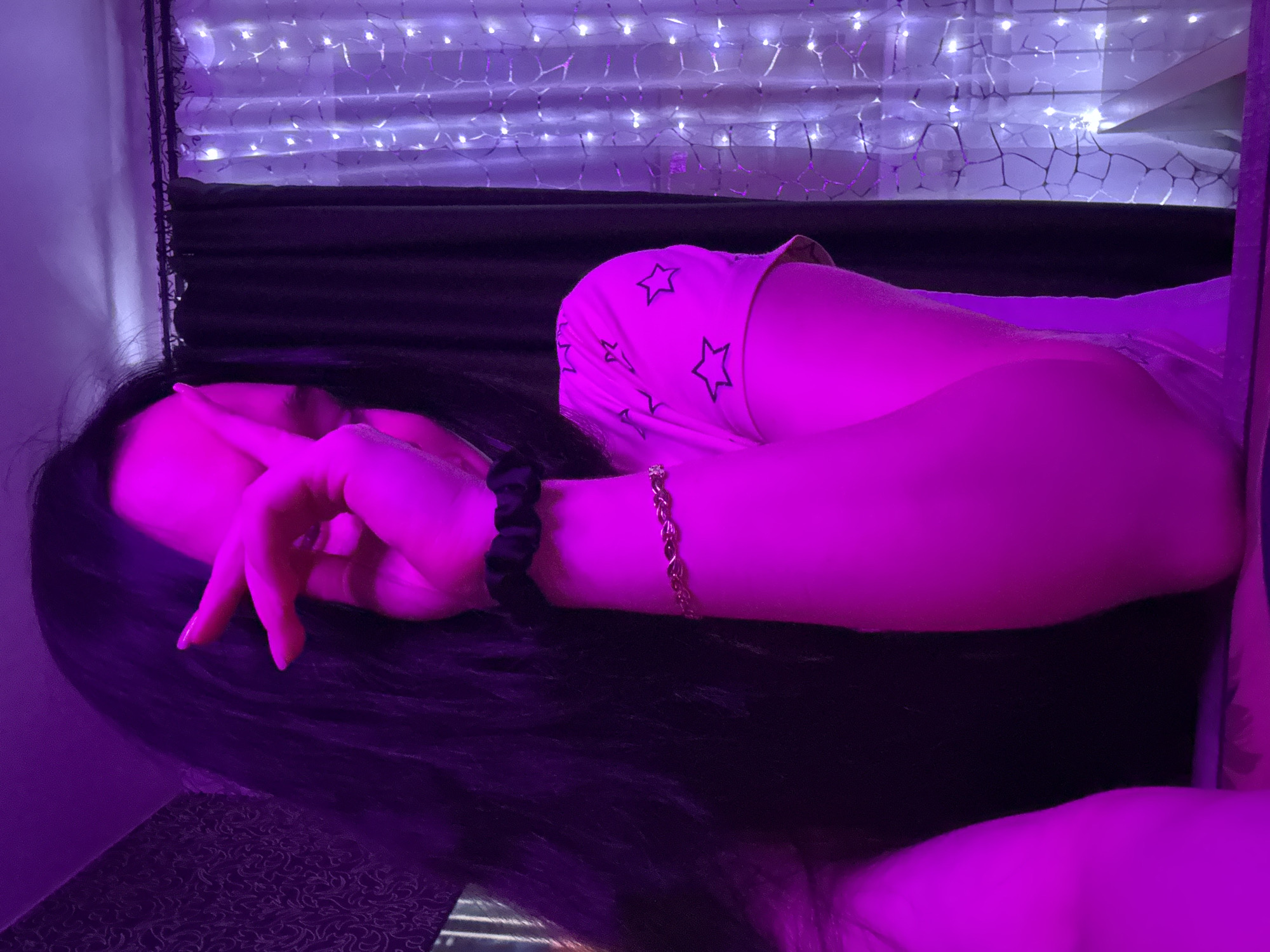 An individual with long, dark hair, illuminated by purple lighting, is visible in a close-up shot. The person is holding their hand up, covering their face partially. They are wearing a light-colored shirt with a star pattern on the sleeve and a thin gold bracelet on their wrist. The background includes dark curtains and sheer curtains decorated with string lights. The overall scene suggests an indoor setting, possibly a bedroom, with a decorative and somewhat intimate atmosphere. There is no identifiable information about the location being Novosibirsk, Russia.