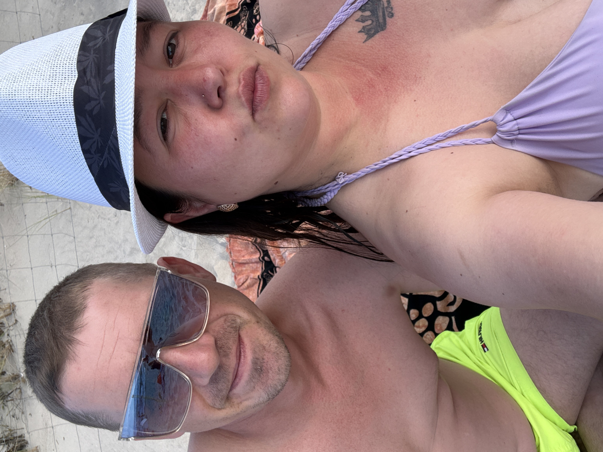 This is a selfie taken by two individuals on a sandy beach, likely in Międzyzdroje, Poland, given the context. The man on the left is wearing sunglasses with blue lenses and neon yellow swim trunks. He has a short haircut and is showing a slight smile. The woman on the right is wearing a white hat with a black band and a light purple bikini top. She has a nose ring and her neck and chest show signs of sunburn. A tattoo of a crown is visible on her chest. They are both seated, possibly on a towel or blanket. The background is blurred but suggests a beach environment with sand and some vegetation.