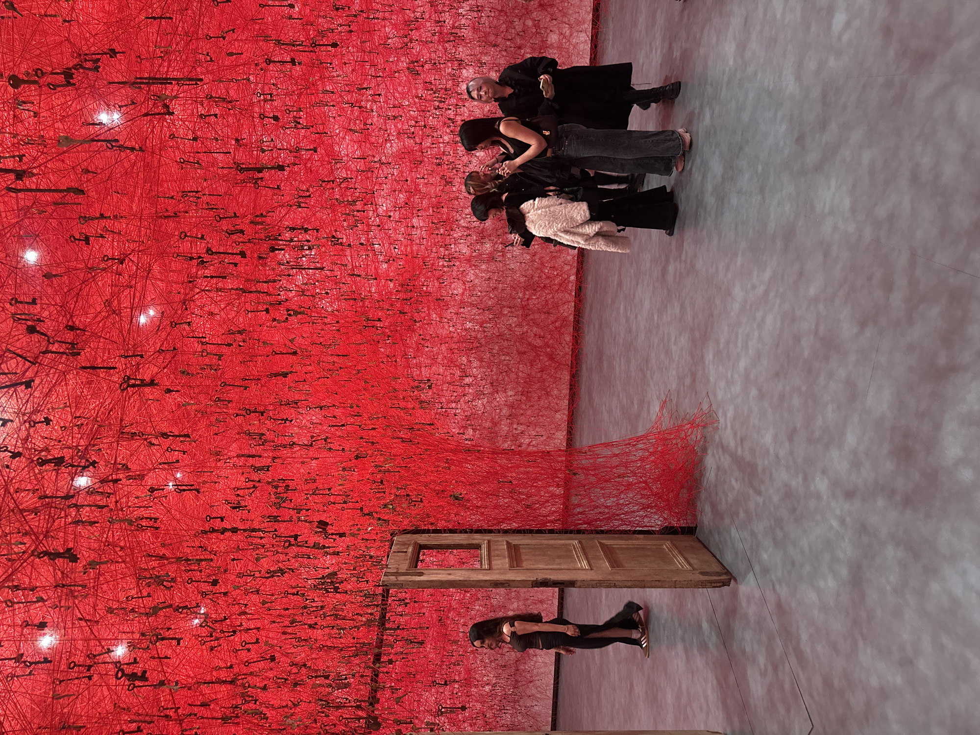 The vertically oriented image captures an immersive art installation within a gallery or museum, located in London, United Kingdom. The left side of the frame is dominated by a vast, dense network of bright red threads or yarn, forming an intricate, interwoven web. Hundreds of metallic, antique-style keys are suspended throughout this crimson matrix, appearing to float within the structure. Several bright, circular light sources are visible within the red threads, illuminating the keys and adding depth to the visual experience.
On the right side of the image, a grey, concrete-like floor extends, showing subtle reflections. A group of four individuals stands on this floor, observing the installation. They are dressed in dark, casual clothing; one person with short, light-colored hair looks towards the viewer with a slight smile, while the others are focused on the artwork. One individual in the group wears a distinct white or cream furry jacket.
Towards the lower right corner, an old, distressed wooden door stands upright and partially ajar, seemingly integrated into the artwork. Red threads extend from and around this door, connecting it to the larger red web. An additional individual with long dark hair, wearing dark attire, stands near this door on the grey floor, also looking towards the striking red installation.
The scene depicts visitors engaging with a large-scale, conceptual art piece, recognized as "The Key in the Hand" by artist Chiharu Shiota. The atmosphere conveys a sense of wonder and artistic contemplation. No discernible text is visible in the frame.