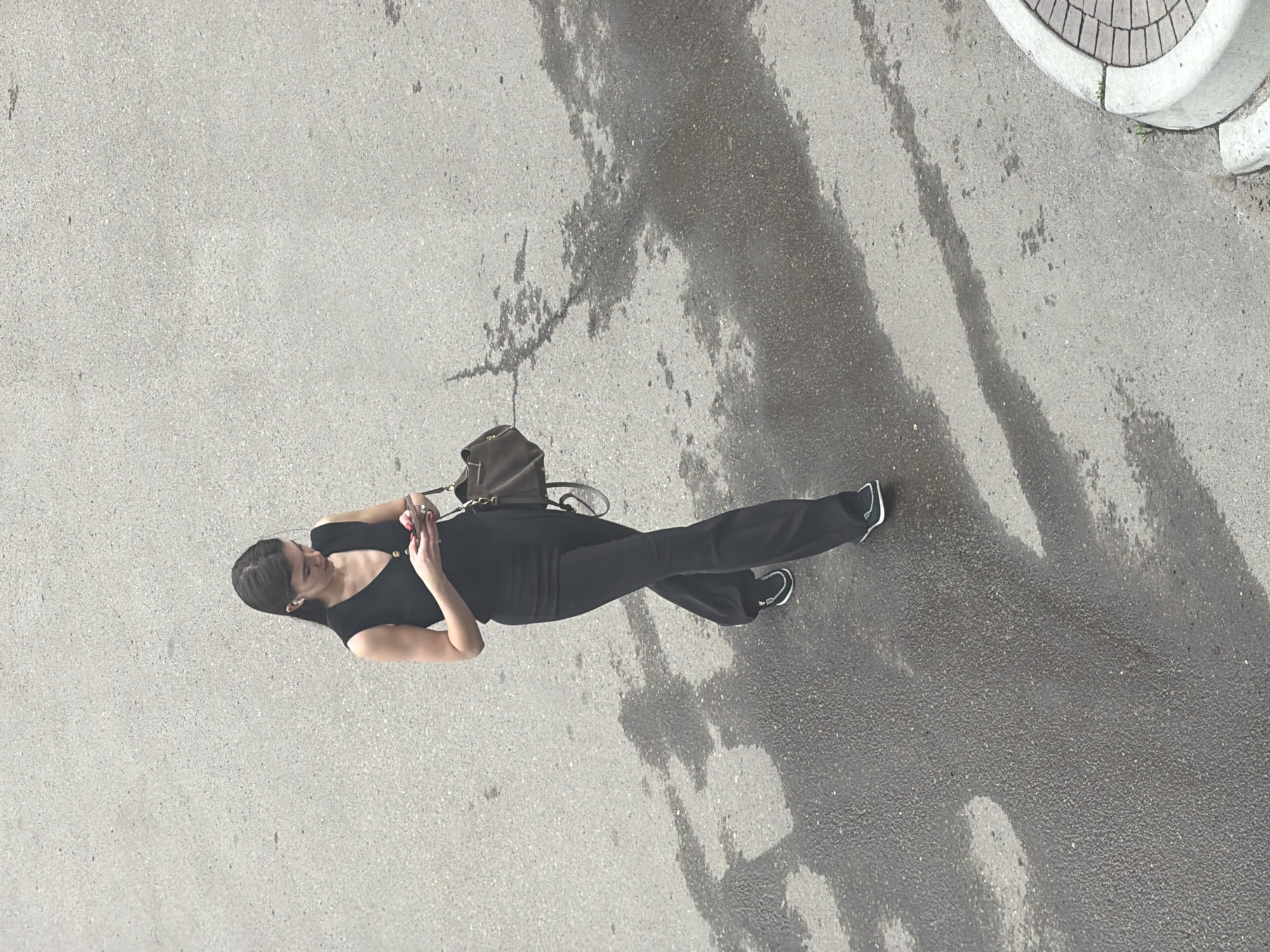A woman in a black outfit and black sneakers walks on a wet asphalt surface. She is wearing a black v-neck top with two buttons, and black flared pants. She has a dark green crossbody bag over her left shoulder. She is holding a smartphone in her right hand. The woman has dark hair and appears to be wearing small white earbuds. The scene is outdoors with a textured asphalt ground that has dark wet patches, indicating recent rain or a spill. In the upper right corner, a portion of a concrete curb with brick paving is visible. The lighting suggests it is daytime, with overcast weather. The image is taken from a high angle looking down at the woman.