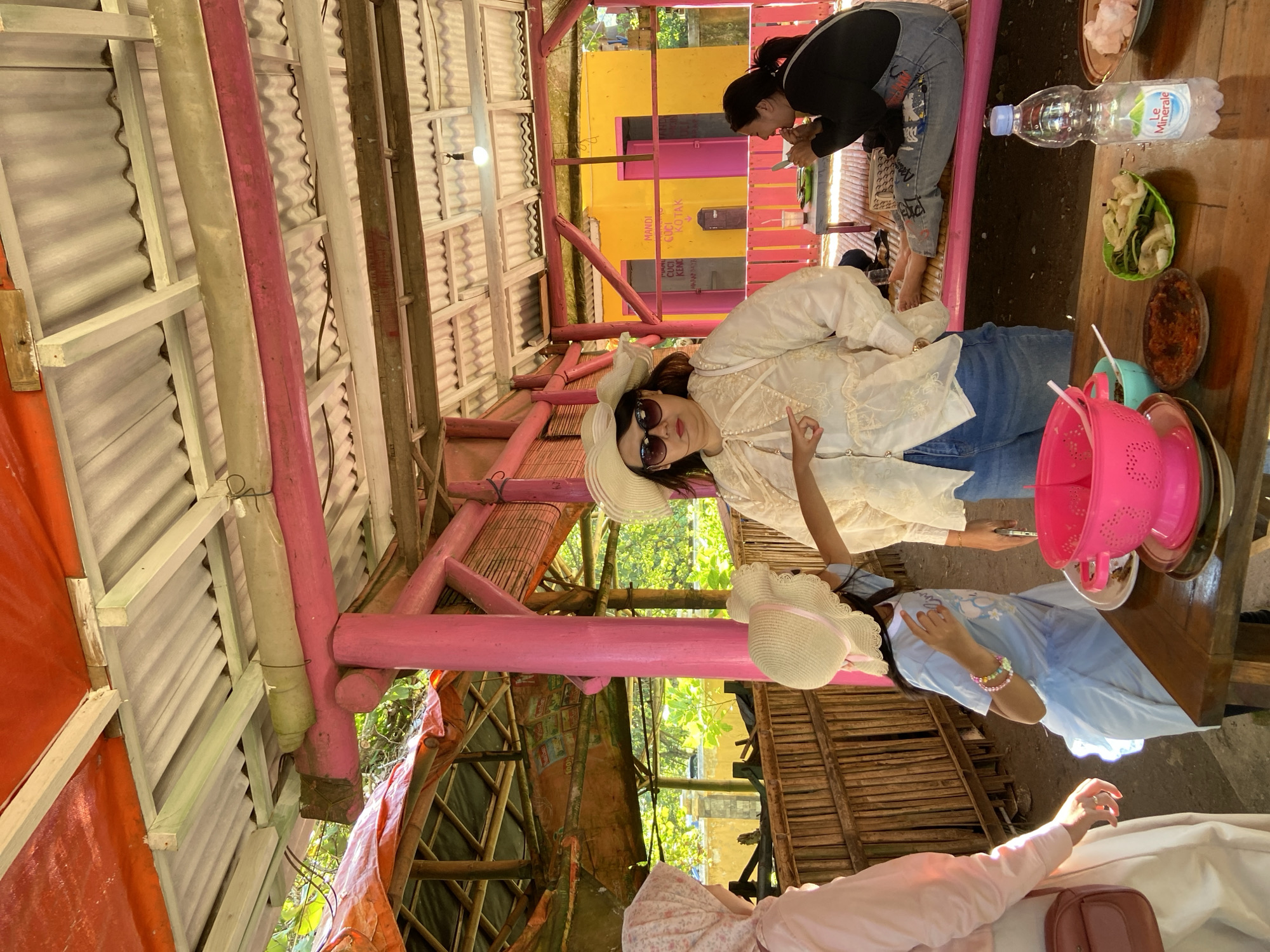 Here is a detailed description of the image:
The image shows an outdoor dining scene in Unknown, Indonesia, possibly within a restaurant or a casual dining setting. The camera appears to be positioned at an angle, providing an overhead view of the scene.
The primary subjects are several individuals seated at a wooden table. A woman in a white blouse and blue jeans is wearing a wide-brimmed hat and sunglasses; she is making a hand gesture with her right hand. Next to her is another individual in a blue dress and straw hat. Another individual partially visible at the bottom edge of the photo is wearing a pink top and appears to be reaching toward the table. In the background, another woman is visible, wearing jeans and a dark top, interacting with a meal.
On the wooden table, there is a collection of food items, including dishes with sauces, vegetables, and what appears to be rice. A bottle of water is also visible, along with pink plastic colander.
The setting appears to be an open structure with a corrugated iron roof supported by brightly colored, mostly pink, wooden beams and posts. A wall with signage in Indonesian language is in the background. The overall environment has a casual and lively atmosphere, suggesting a social gathering or meal shared among friends or family.