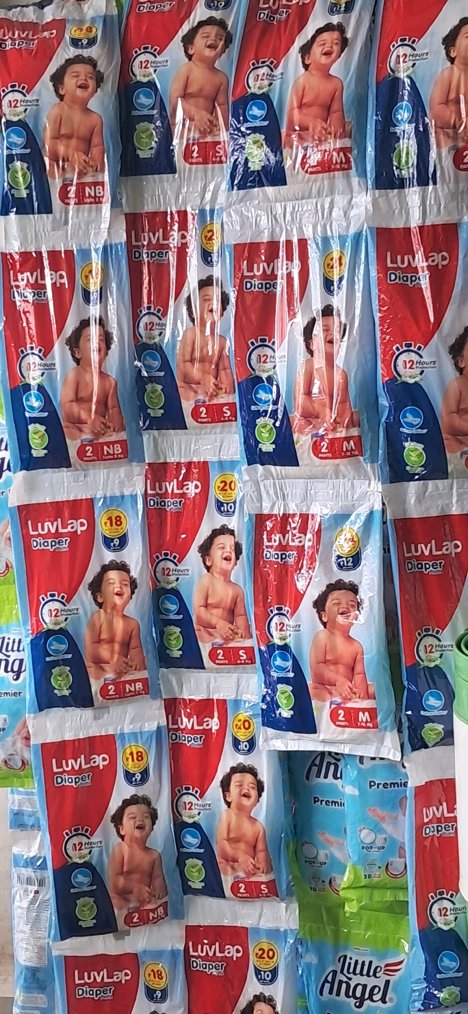 The image shows a wall display of multiple packs of baby diapers. The diapers are packaged in brightly colored wrappers, primarily red, blue, and white, with images of babies on them. The brand name "LuvLap Diaper" is prominently displayed on many of the packs, along with information like "12 Hours Protection," sizing indicators (NB, S, M), and the number of diapers per pack (e.g., "2"). Some packs also feature circular icons with numbers like "18" and "9," and "20" and "10." The diapers are arranged in vertical rows, suggesting they are stacked or hanging for display in a retail setting. The overall environment appears to be indoors, likely a store or market. The location is specified as Rayachoty, India.
