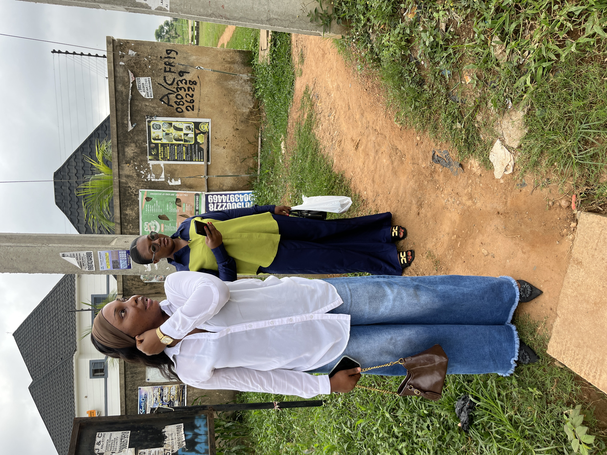 Two Black women stand on a dirt path in a grassy, urban area in Abuja, Nigeria. The woman in the foreground is wearing a white button-down shirt, wide-leg blue jeans, and black pointed shoes. She is holding a brown purse in her left hand and a smartphone in her right. She has a brown headscarf and is looking to her left.
The woman in the background is wearing a green and blue top and dark blue pants. She is holding a smartphone in her right hand and a white plastic bag in her left. She is wearing sunglasses and looking down at her phone.
Behind the women is a concrete wall with several posters and graffiti. There is also a pole with an electric fence on top. In the distance, a modern house with a dark roof is visible. The overall scene suggests a casual outdoor setting in a residential or commercial area of Abuja.
