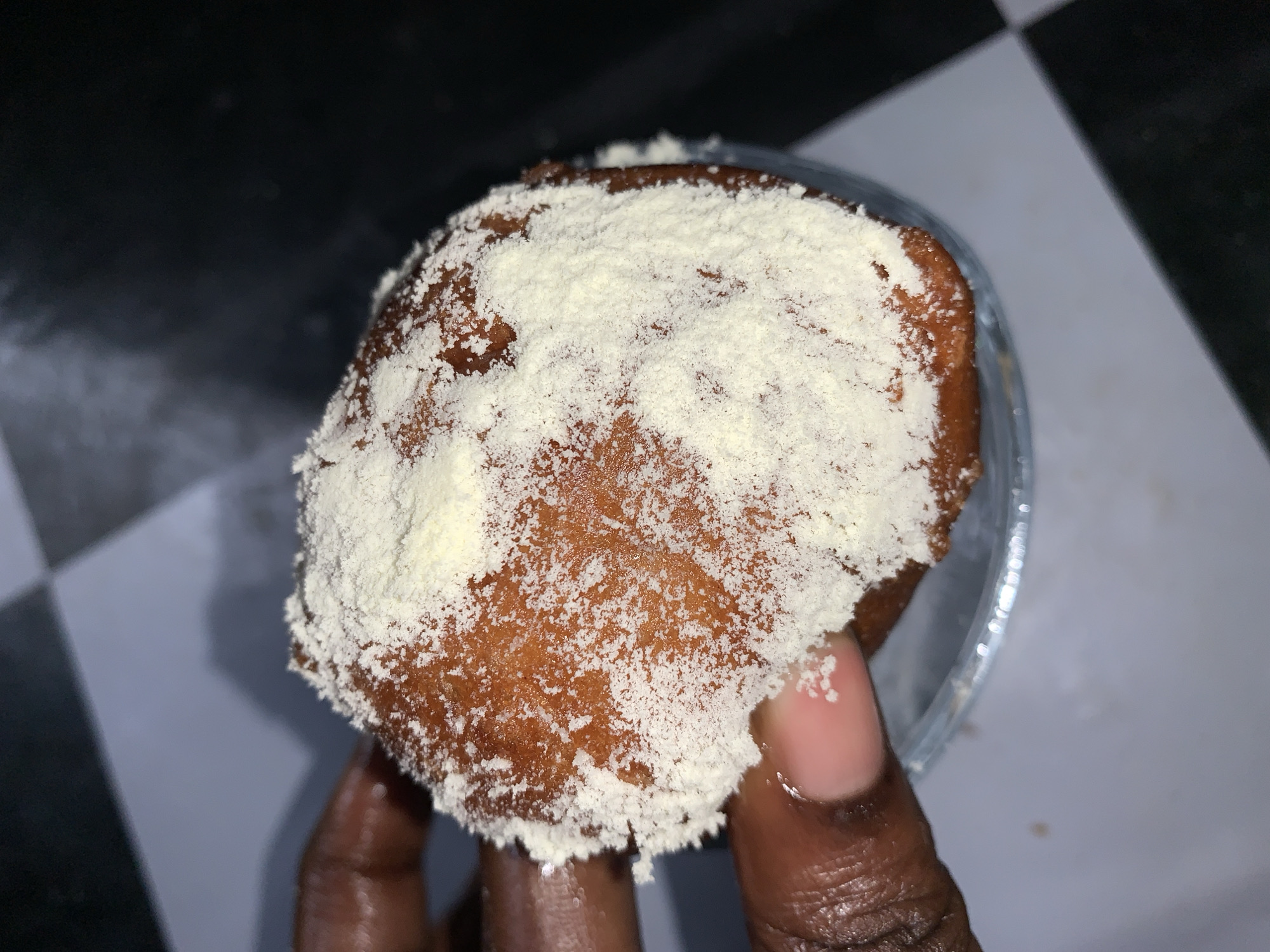 An overhead, close-up shot captures a hand holding a fried dough pastry that is dusted with a white powder. The pastry is golden brown, round, and slightly flattened. The white powder appears to be finely ground. The hand is dark-skinned, with visible fingerprints and nails. The background is a black and white checkered surface, suggesting a countertop or floor. The lighting is dim, creating shadows and highlighting the texture of the pastry and the powder. The image does not provide any information about the location in Rafin Par, Nigeria.