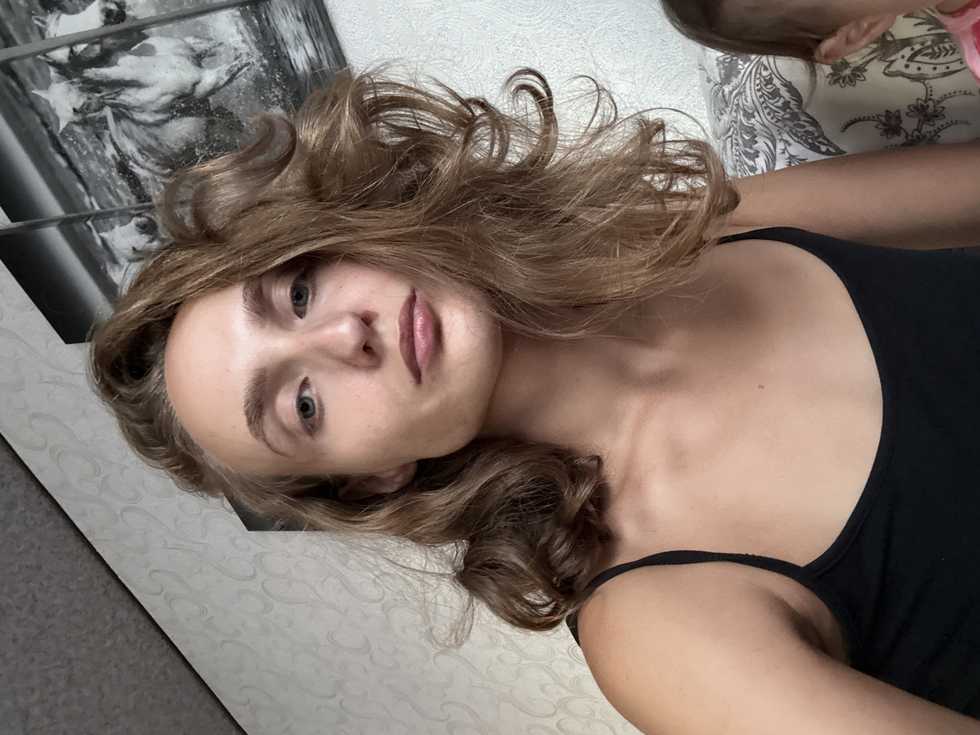The image is a close-up, selfie-style portrait of a young Caucasian woman with light brown, wavy hair. She is wearing a black tank top and is positioned in the foreground. Her gaze is directed towards the camera. In the background, a wall with a patterned texture is visible, along with a framed artwork depicting horses. To the right of the woman, a portion of a child's face and shoulder, wearing a pink garment, is also visible. The overall scene appears to be indoors, likely a residential setting. There is no identifiable information indicating the location in Kuyeda, Russia.