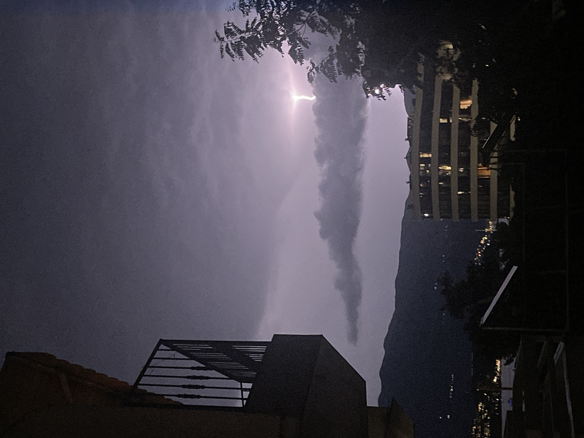 A horizontal lightning bolt illuminates the sky above a cityscape at night, with the mountains of Budva, Montenegro, visible in the background. A balcony railing juts into the frame from the left foreground, while a multi-story building with illuminated windows stands to the right of the frame, partially obscured by dark trees. The sky is filled with heavy, dark clouds, indicating stormy weather.