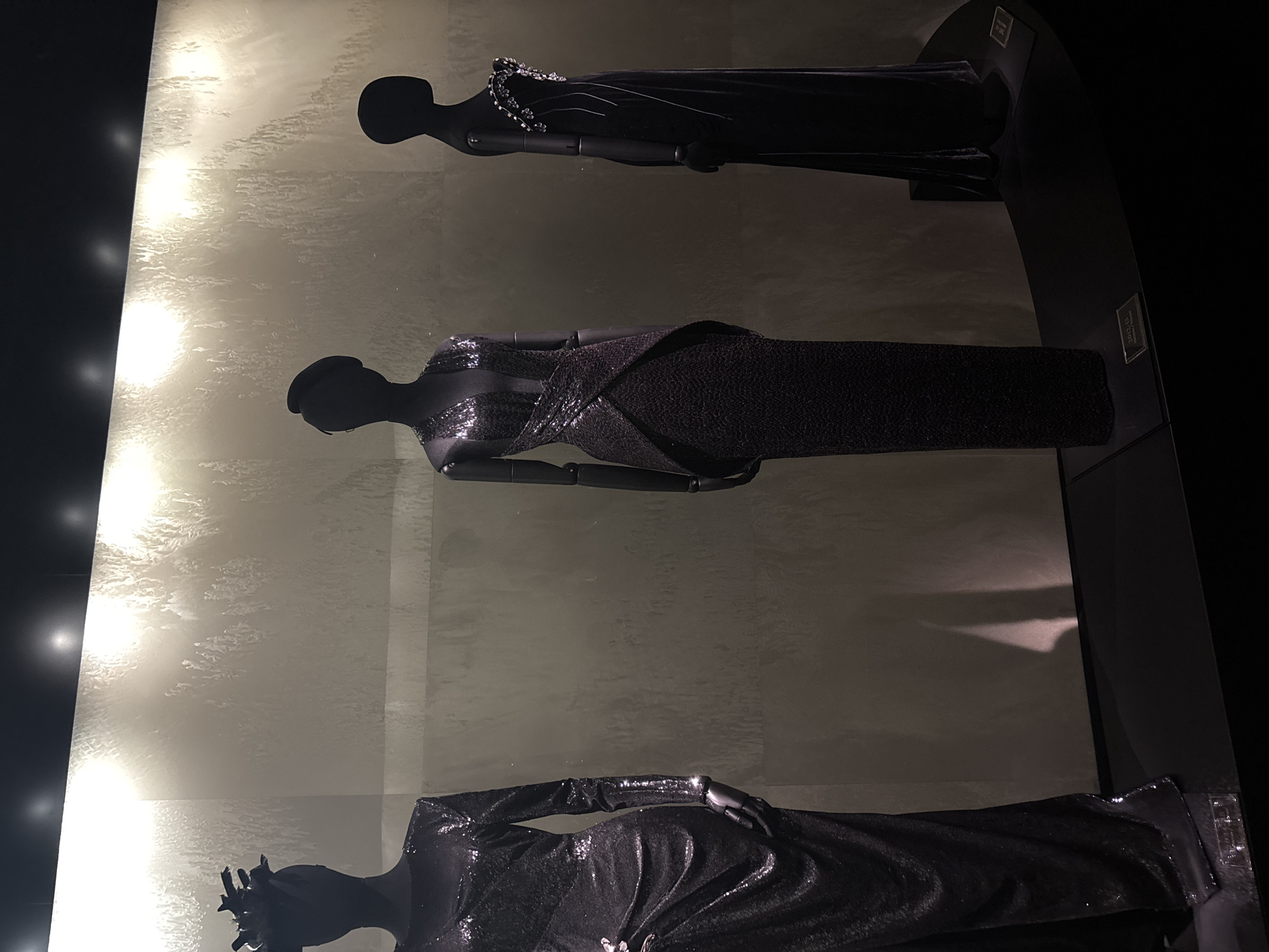 Three mannequins are displayed in a dimly lit exhibition space. The mannequins are dark, silhouetted figures wearing elaborate, floor-length gowns. The background is a textured, light-colored wall with dramatic lighting that creates a shimmering effect. The mannequins are positioned on a dark, curved platform. The overall aesthetic suggests a fashion exhibition, possibly showcasing haute couture. The scene appears to be an indoor exhibit, with spotlights focused on the display. No people are visible in the image.