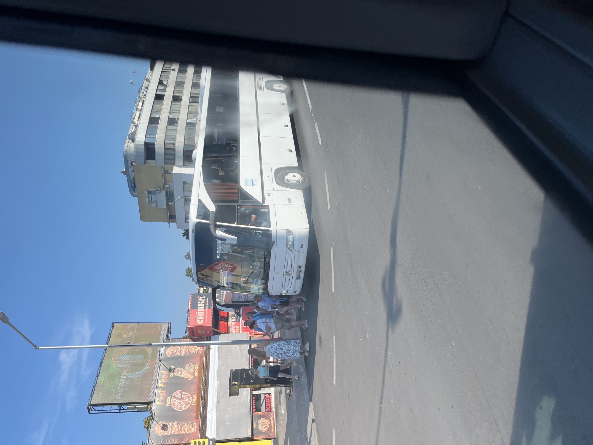 A white Mercedes-Benz tour bus is parked on a street in Nesebar, Bulgaria, next to a sidewalk with a group of tourists. The bus is emitting a significant amount of white exhaust smoke from its rear. Behind the bus, there are several fast-food stands with advertisements for "Fast Food," "Pizza," and "Döner." A tall streetlight stands to the left of the bus. In the background, there is a modern apartment building with balconies and satellite dishes. The sky is clear and blue, indicating a sunny day. The scene appears to be a common occurrence at a tourist bus stop or a parking area near food establishments.