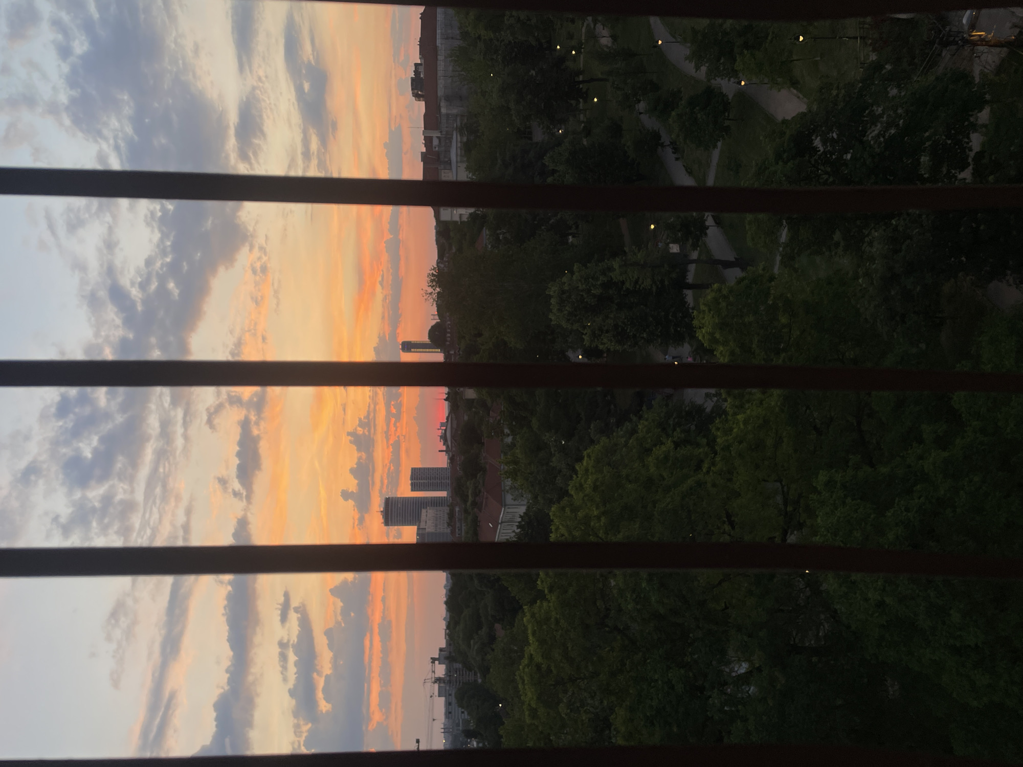 The image shows a view through horizontal window blinds of an orange and pink sunset over an urban landscape in Vracar Urban Municipality, Serbia. To the left of the image the sky is visible between the slats of the window blinds, including orange, pink, and grey clouds. Some buildings are visible in the middle. To the right is a park, and some city streetlights are visible in the park. There are no people visible.