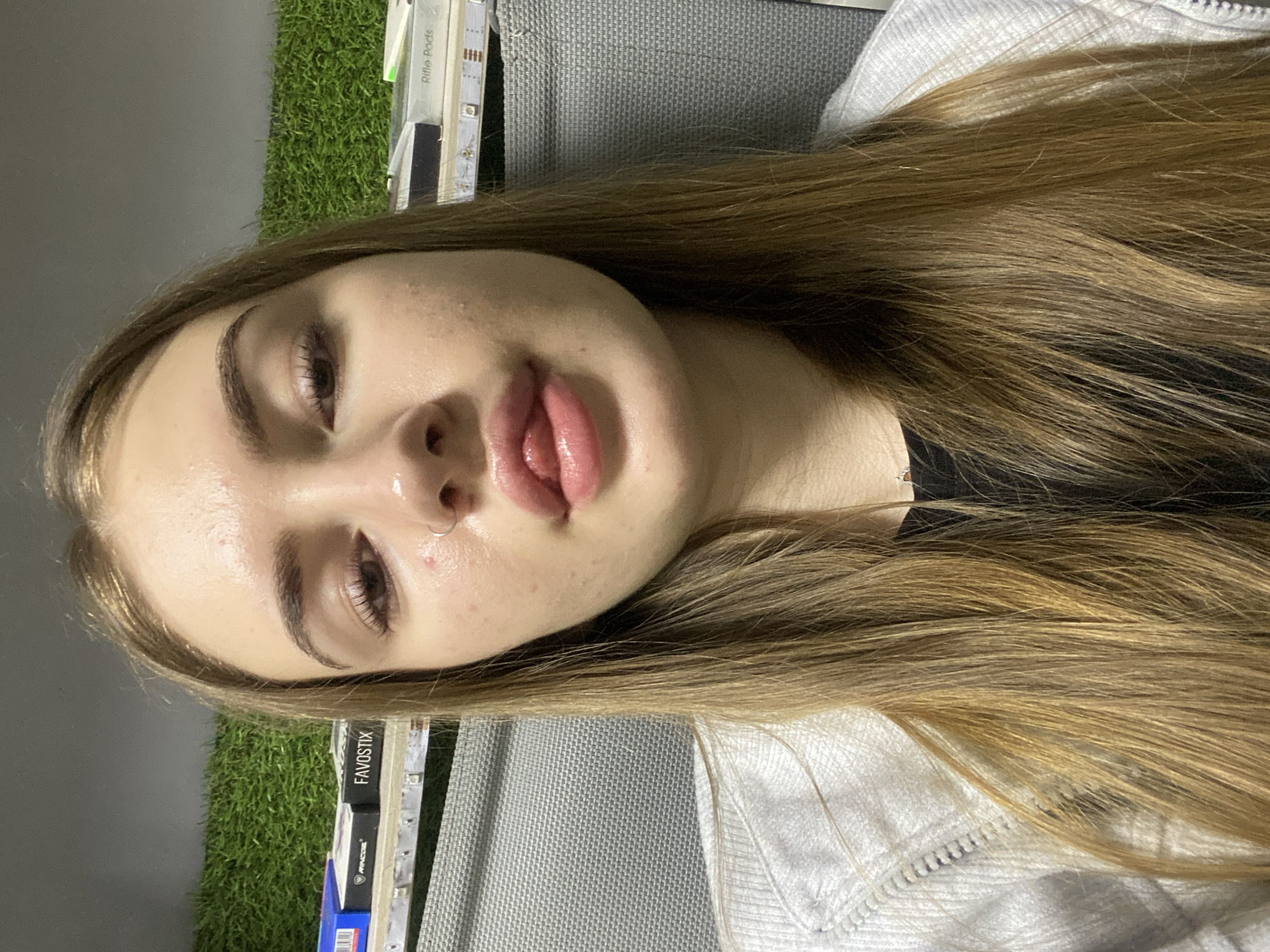 The image depicts a young woman in an indoor setting in the city of Дьяково, Russia. She is shown from the chest up, looking directly at the camera in what appears to be a selfie. Her light brown hair is long and straight, parted in the middle. She has a small silver hoop nose ring on her left nostril. Her mouth is slightly open, with her tongue protruding upwards towards her upper lip, which is covered in a glossy substance. Her skin appears dewy, with some visible pores and minor blemishes on her forehead and cheeks. She is wearing a white zippered jacket or hoodie over a black inner garment.
The background features a vertical surface covered in artificial green grass, suggesting a decorative wall. Below this, a light-colored shelf or ledge is visible, adorned with a strip of illuminated LED lights. On the left side of the shelf, several boxes are stacked, including one prominently displaying "FAVOSTIX" and another labeled "AVCOS". A blue box is partially visible further left. On the right side of the shelf, similar white boxes, one partially legible as "Rifle Pods", are also stacked. To the right of the woman, a grey, textured surface, possibly upholstery or a panel, is visible.