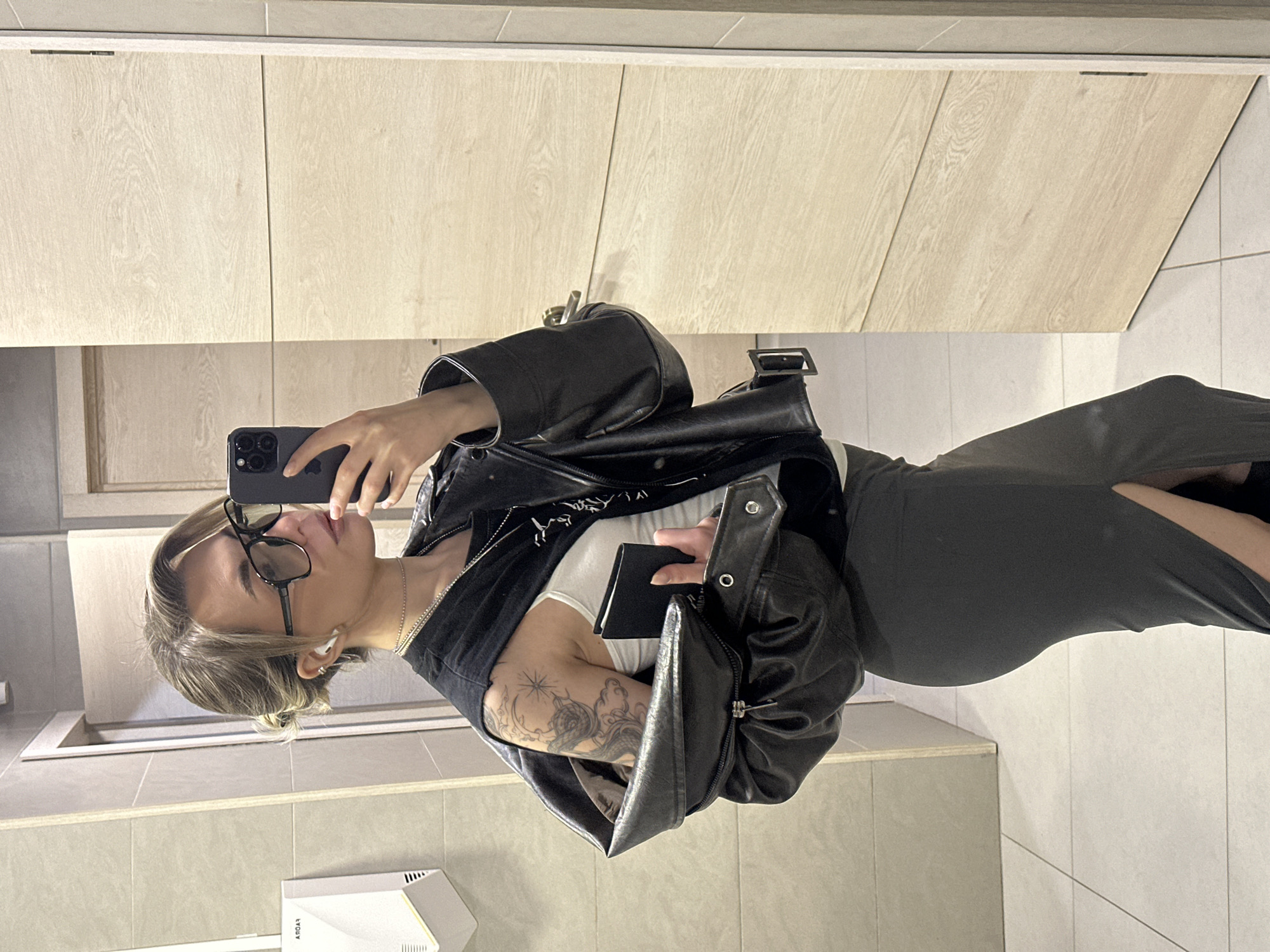 A woman is taking a selfie in a bathroom. She is wearing glasses, a black jacket, a white top, and a grey dress with a slit. Her left arm has a tattoo. She is holding a smartphone in each hand. The bathroom has light wood cabinets, beige tiles, and a white wall-mounted device. There is no evidence of the location being Khimki, Russia.