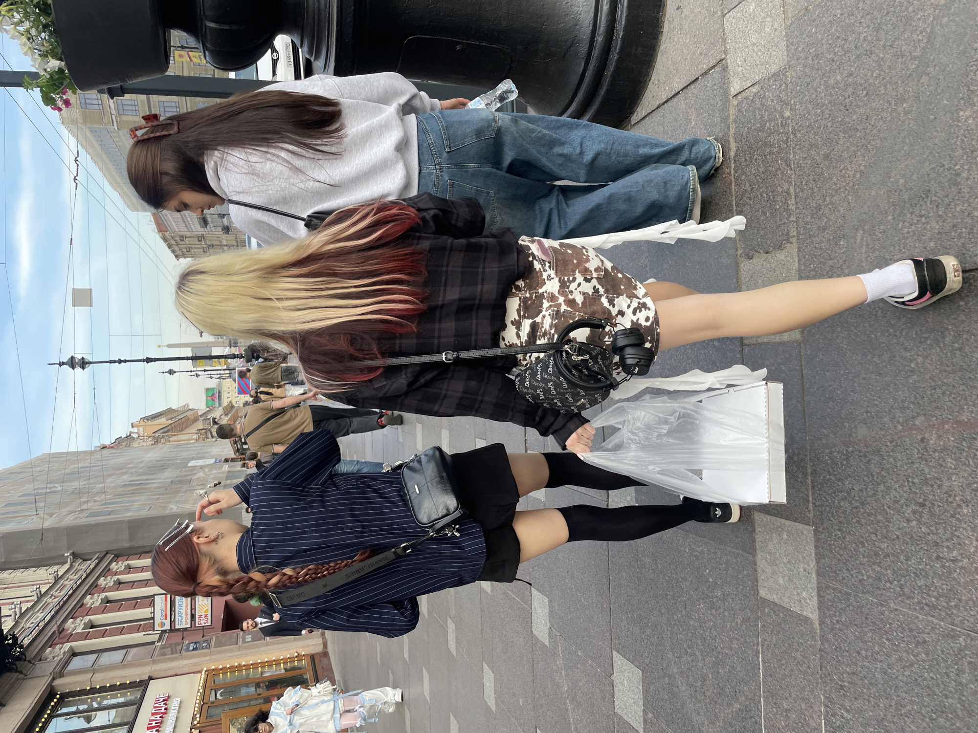The image depicts a street scene in what appears to be Saint Petersburg, Russia, identified by architectural details and Cyrillic script on signage. Three young women are the main subjects, positioned near what could be a street lamp or a decorative element.
The woman closest to the camera wears a navy pinstripe shirt, a black skirt, knee-high black socks, and black shoes. She has a crossbody black bag and a braid with red highlights in her hair. Her face is partially visible; she is holding a finger near her face and has hair clips in her hair.
The woman in the middle has blonde hair with red undertones, a plaid jacket, and a cow print skirt. She carries a black purse and has headphones around her neck. She also carries a white rectangular box in a plastic bag.
The third woman, positioned further away, is dressed in a grey sweatshirt and jeans.
The background features an urban setting with buildings, architectural details like street lamps, and other people walking on the street. Signage in Cyrillic script can be observed above some shopfronts.