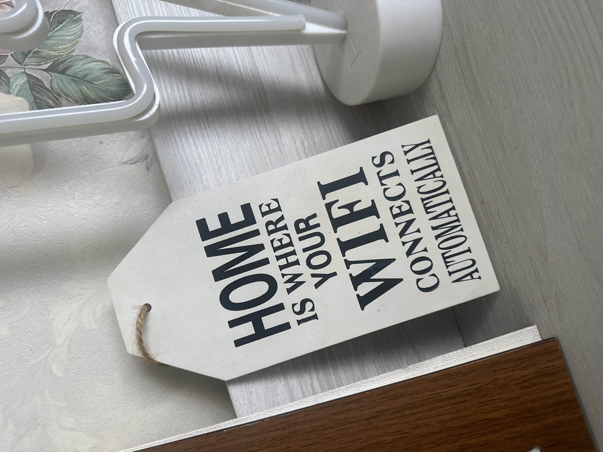 The image shows a tag with the text "HOME IS WHERE YOUR WIFI CONNECTS AUTOMATICALLY". The tag is off-white with black lettering, and there's a string hanging from the top. The tag is resting on a wooden surface next to a white metal structure and another off-white surface with a floral pattern. The background consists of a light gray, smooth surface. The location information is not discernible from the image.