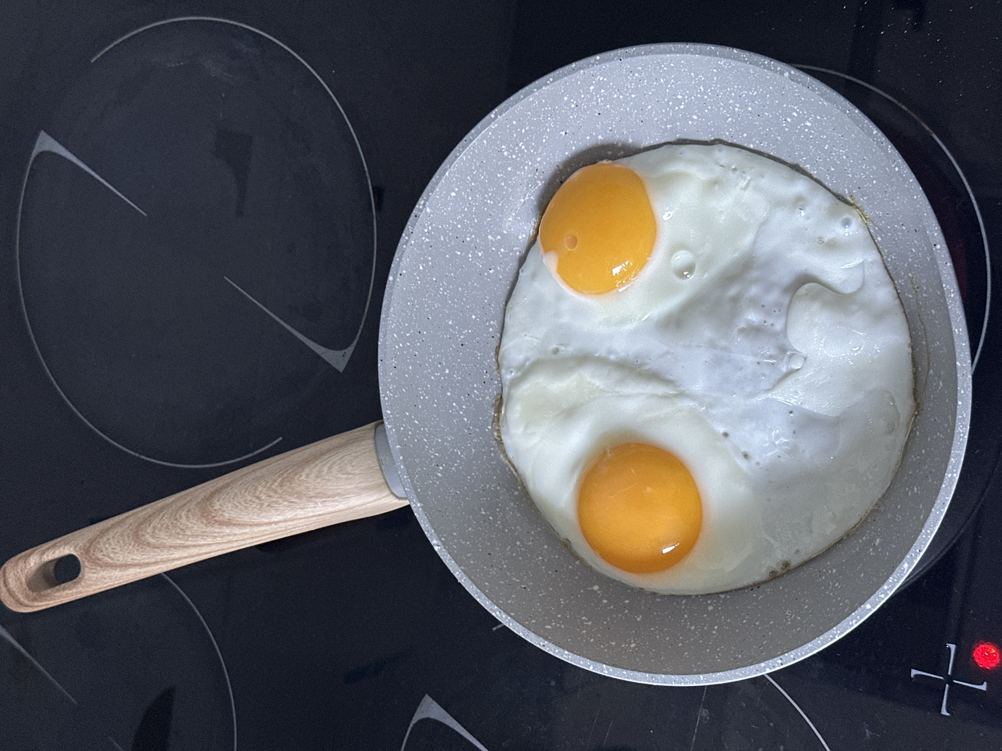A fried egg is cooking in a gray speckled pan with a light brown wooden handle on a black induction stovetop. The pan contains two sunny-side-up eggs with bright orange yolks and cooked white egg whites. The stovetop has visible circular heating elements and a red indicator light glowing on the lower left. The scene appears to be an indoor kitchen setting, likely in Moscow, Russia, given the question's context.