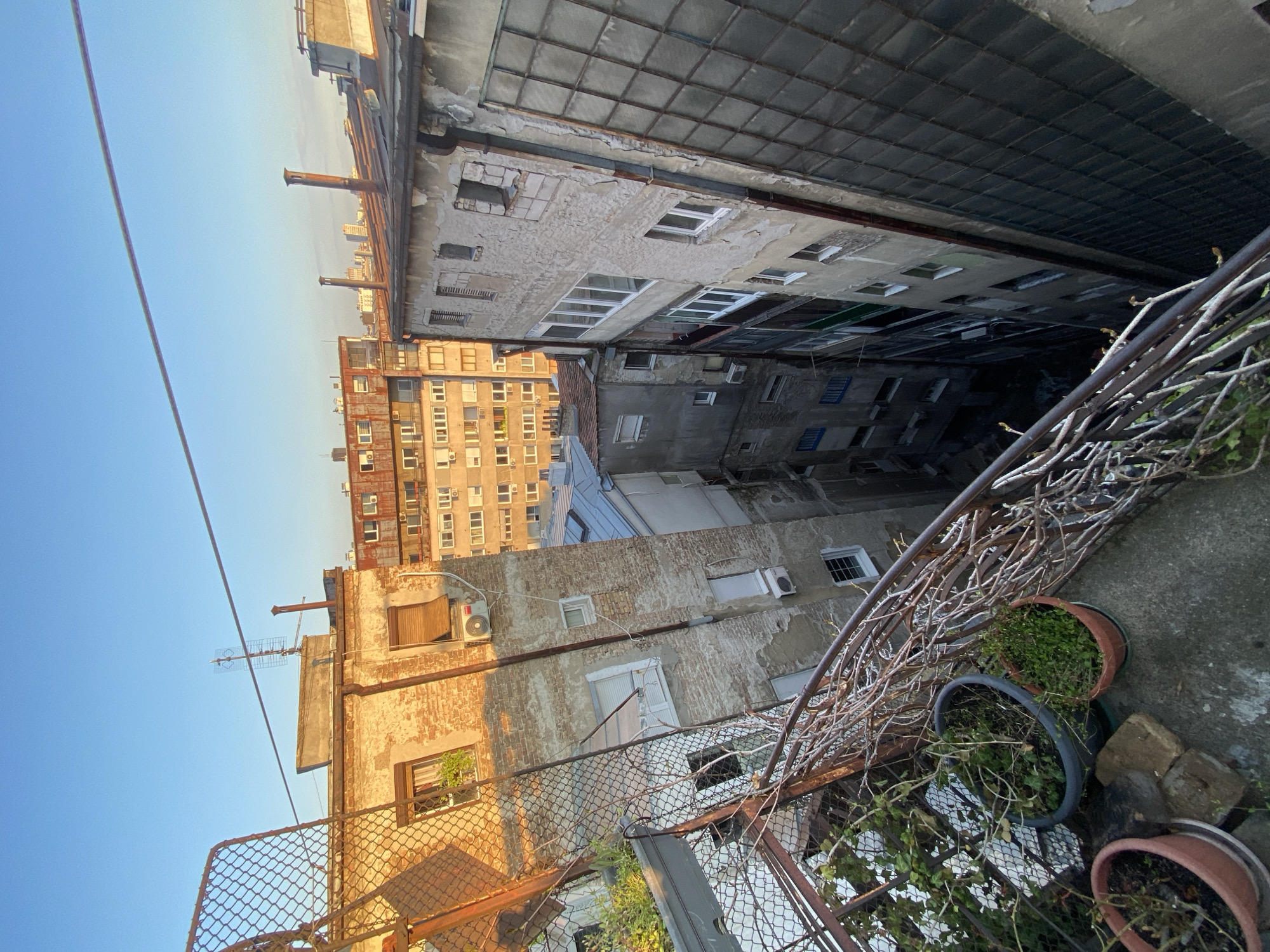 An upward, slightly angled shot looking into a narrow alleyway or space between tall, aged buildings in Belgrade, Serbia. The buildings are a mix of brick and concrete, with visible wear and tear, including peeling paint, crumbling facades, and a variety of window styles. Some windows are open, while others are covered with curtains or shutters. Air conditioning units are mounted on some of the buildings. A chain-link fence and a metal railing with climbing plants and potted plants are visible in the foreground, suggesting a balcony or rooftop area. The sky is visible at the top of the frame, indicating the open space above the buildings. The scene conveys a sense of urban decay and the confined nature of living in a densely built environment. No people or specific actions are visible.