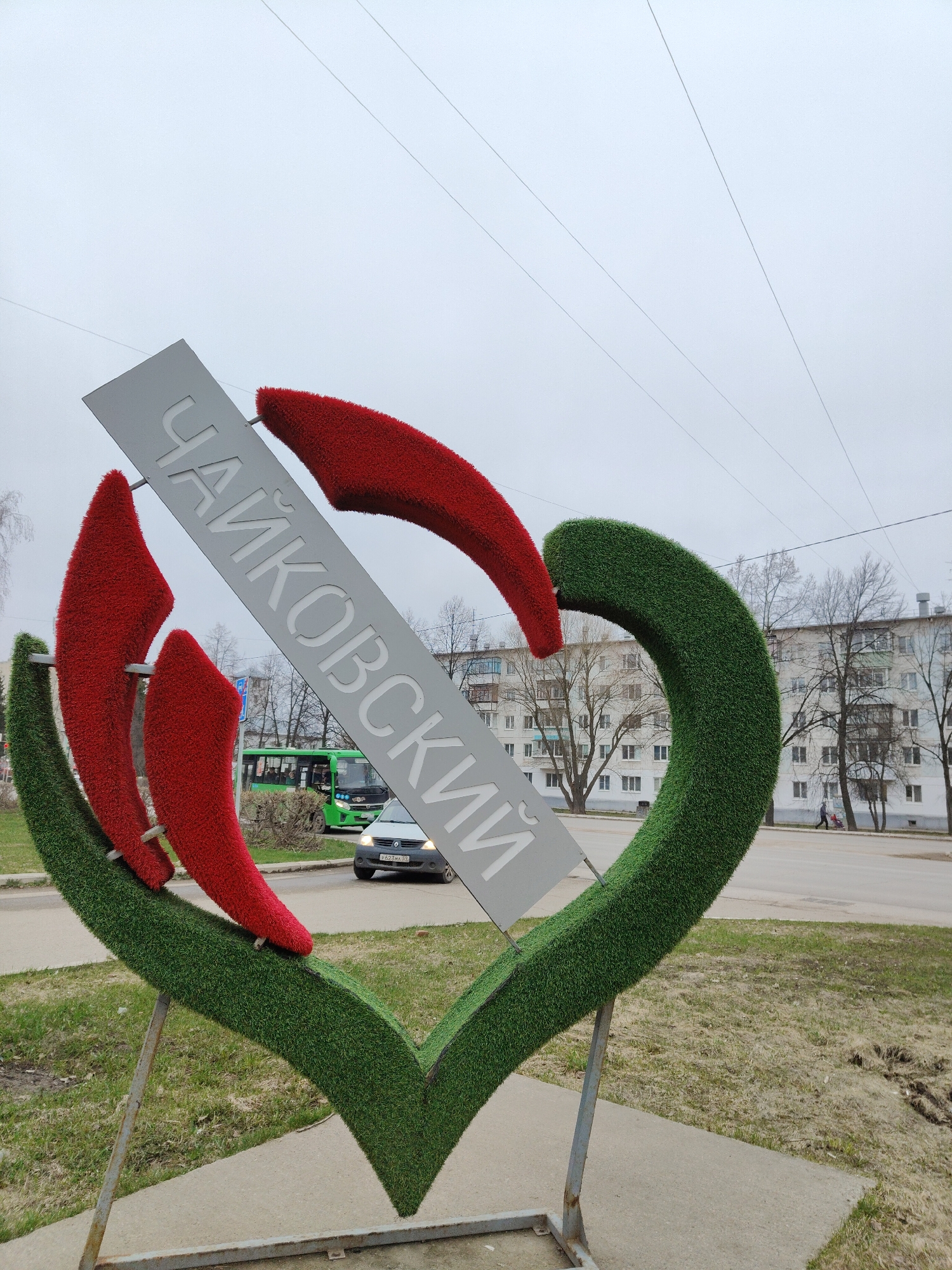 The image displays a large, decorative monument featuring the word "ЧАЙКОВСКИЙ" (CHAIKOVSKY) in Russian Cyrillic on a tilted, gray rectangular panel. This panel is set within a prominent heart-shaped structure, which is covered in green artificial turf. Inside the green heart, two red, stylized, flame-like shapes, also made of artificial turf, provide a contrasting pop of color. The monument is supported by metal poles anchored in a concrete base, with patches of dry grass and dirt on the surrounding ground.
In the background, an urban street scene is visible under an overcast, light gray sky. A paved road runs horizontally across the mid-ground, with a white car (likely a Renault Logan) showing the license plate "P623MA 59" and a green city bus. Beyond the street, a sidewalk has a person walking with a stroller. Behind the street and sidewalk are several multi-story apartment buildings, characterized by their light-colored facades and numerous windows. Bare, deciduous trees are scattered between the buildings and the street. Overhead power lines crisscross the sky. The activity depicted is general urban movement, with vehicles on the road and a pedestrian on the sidewalk. The time of day is daytime, with muted light indicating an overcast weather condition. The Cyrillic text and the Russian license plate ("59" for Perm Krai) indicate the location is likely the city of Tchaikovsky in Russia.