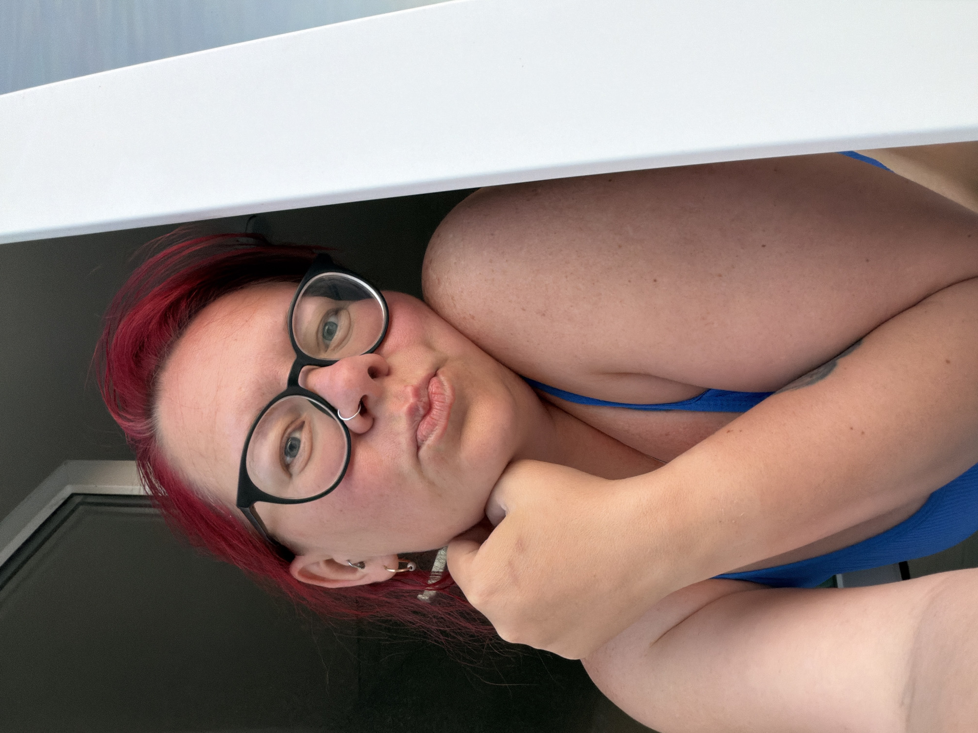 A person with red hair, wearing glasses and a blue top, is photographed indoors from an unusual angle, appearing to be partially under a white surface (possibly a shelf or table). They are holding their chin with their hand. The location is not determinable from the image.