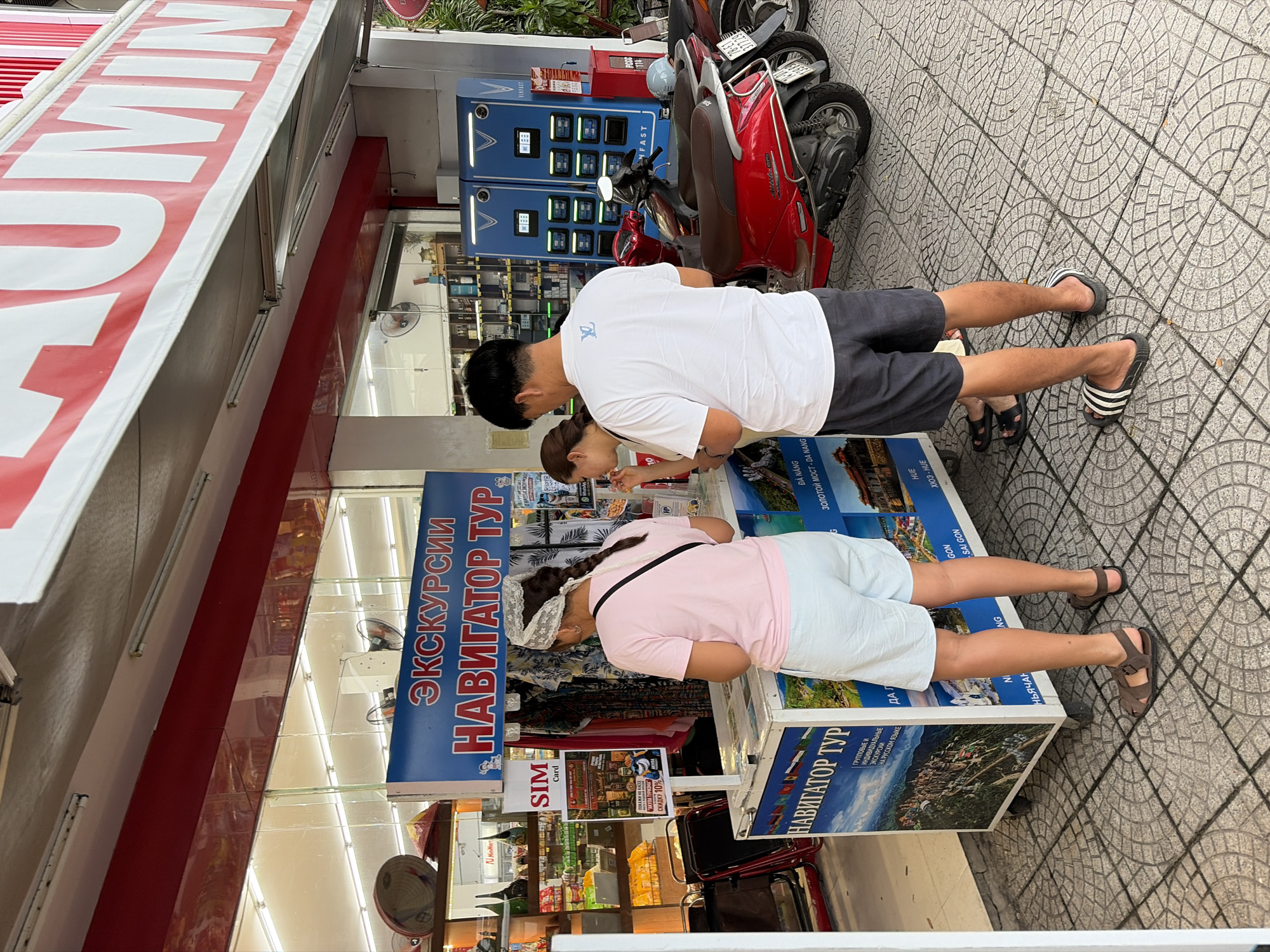 The image shows a street scene in Da Nang, Vietnam. In the foreground, three people are standing around a mobile kiosk. The kiosk displays information about tours and has the words "НАВИГАТОР ТУР" (Navigator Tour) written on it in Cyrillic. One person, a woman with dark hair and wearing a light pink t-shirt and white shorts, is looking at a flyer on the kiosk. Another person, a man with dark hair wearing a white t-shirt and dark shorts, is standing next to her. A third person, a woman with braided dark hair wearing a white lace head covering and a light pink t-shirt, is looking down at the kiosk.
Behind the kiosk, there are two red scooters parked. To the right of the scooters, there are two large blue charging stations with the VinFast logo. The background shows a store with shelves stocked with various goods. The ground is paved with patterned tiles. A large banner with red and white lettering is visible above the store. The lighting suggests it is daytime.