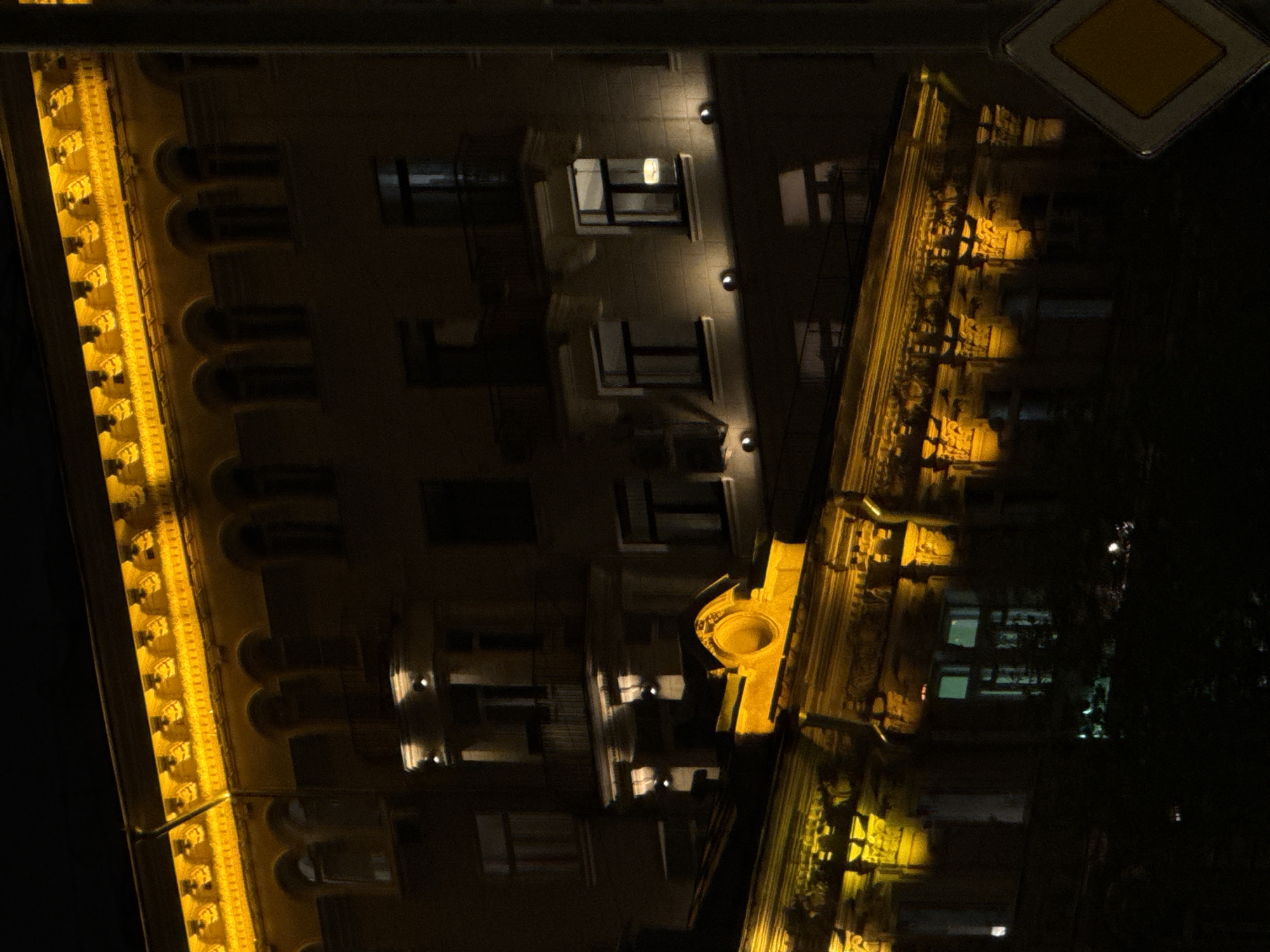 The image shows a section of a building at night in Moscow, Russia, with a street sign visible at the top. The building has multiple windows, some of which are illuminated from the inside. Ornate architectural details line the edge of the building. The scene is mostly dark, with light emanating from the building and the street sign.