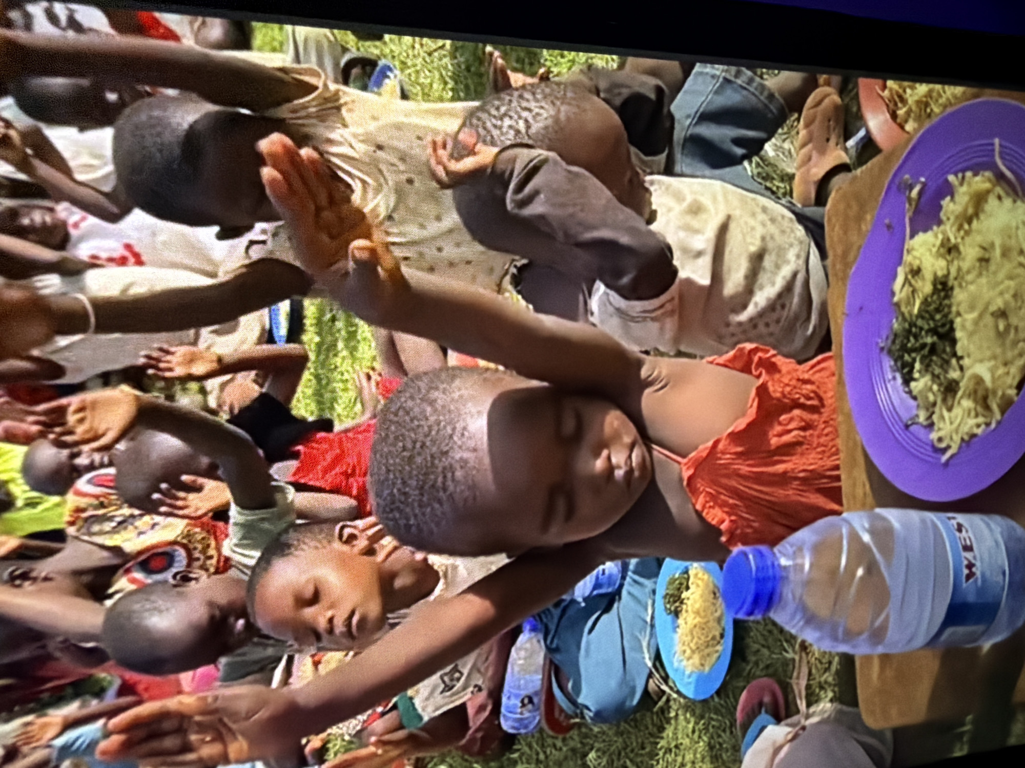 The image shows a group of children outdoors, with their arms raised in the air. The children appear to be of African descent and are dressed in casual clothing. Some of the children have their eyes closed, suggesting they may be praying or singing. In the foreground, there are plates of food, a bottle of water, and some colorful sandals. The ground is covered in grass. The overall scene suggests a gathering or event, possibly a religious service or a meal.