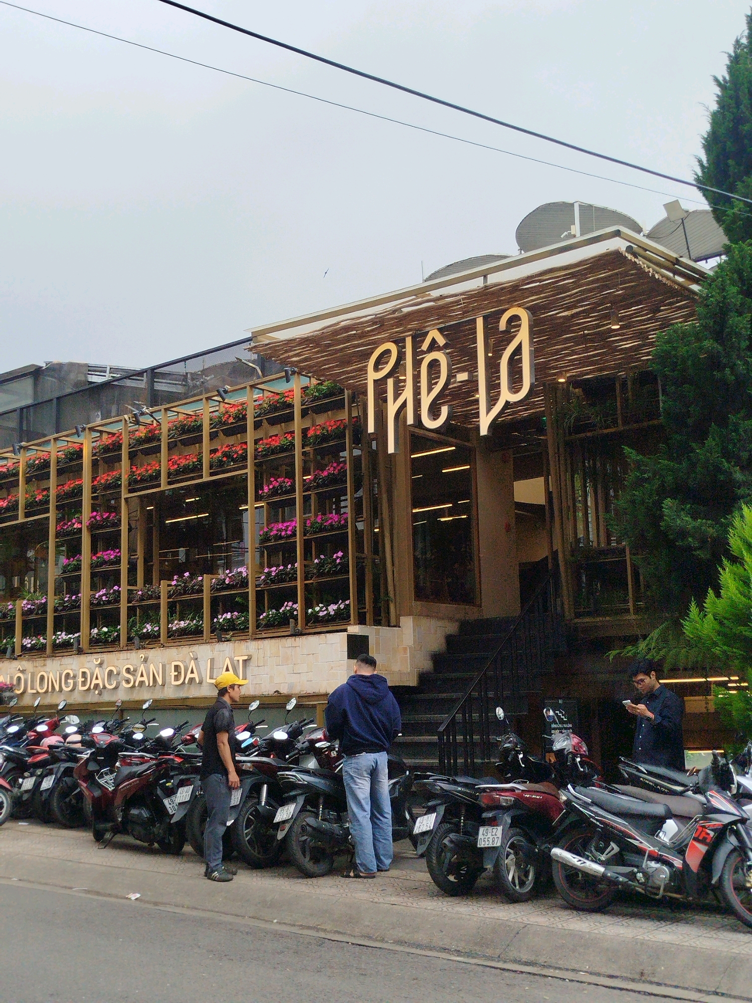 The image shows the exterior of a building in Da Lat, Vietnam, with a distinctive facade featuring vertical shelves filled with flowering plants. A large, illuminated sign above the entrance reads "Phê-la." In front of the building, a row of parked motorcycles lines the street, with several people standing near them. One person in a yellow cap is facing the motorcycles, while another person in a blue hoodie stands with their back to the viewer. A third person, wearing glasses and a dark shirt, is looking at their phone while standing near a motorcycle. The street appears to be paved, and the building's architecture suggests it might be a cafe or a shop. The overall atmosphere is that of a typical urban scene in Da Lat, with its characteristic blend of natural elements and modern infrastructure.