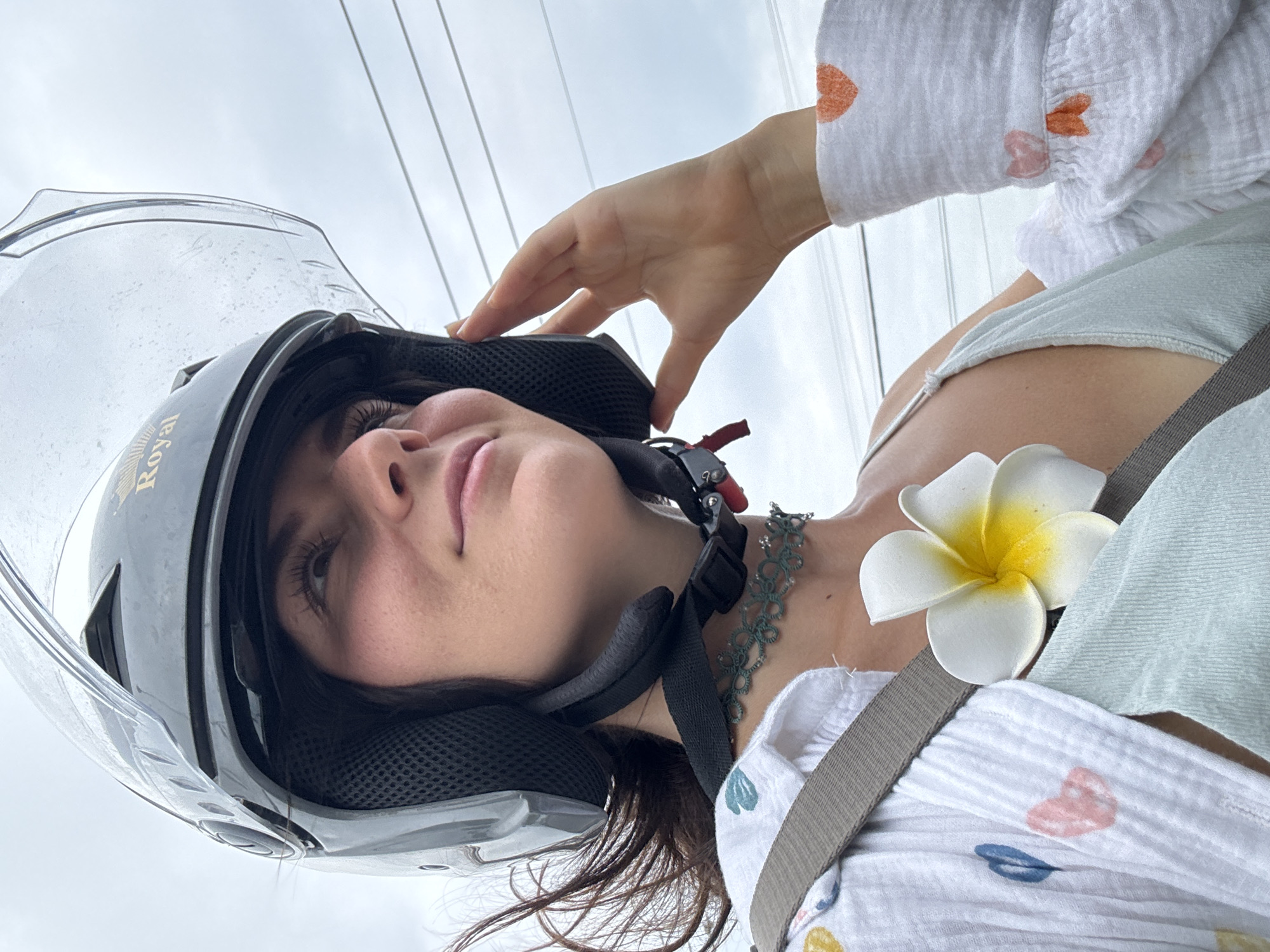 A woman wearing a dark helmet with a clear visor is the subject of a close-up photograph taken in Mũi Né, Vietnam. The helmet is likely for motorcycle or scooter use. Her hand is positioned near the helmet's edge. The woman is also wearing a light-colored top with a pattern of hearts. A white flower with a yellow center is placed near her shoulder. A seat belt strap is visible. Utility lines can be seen against the sky in the background.