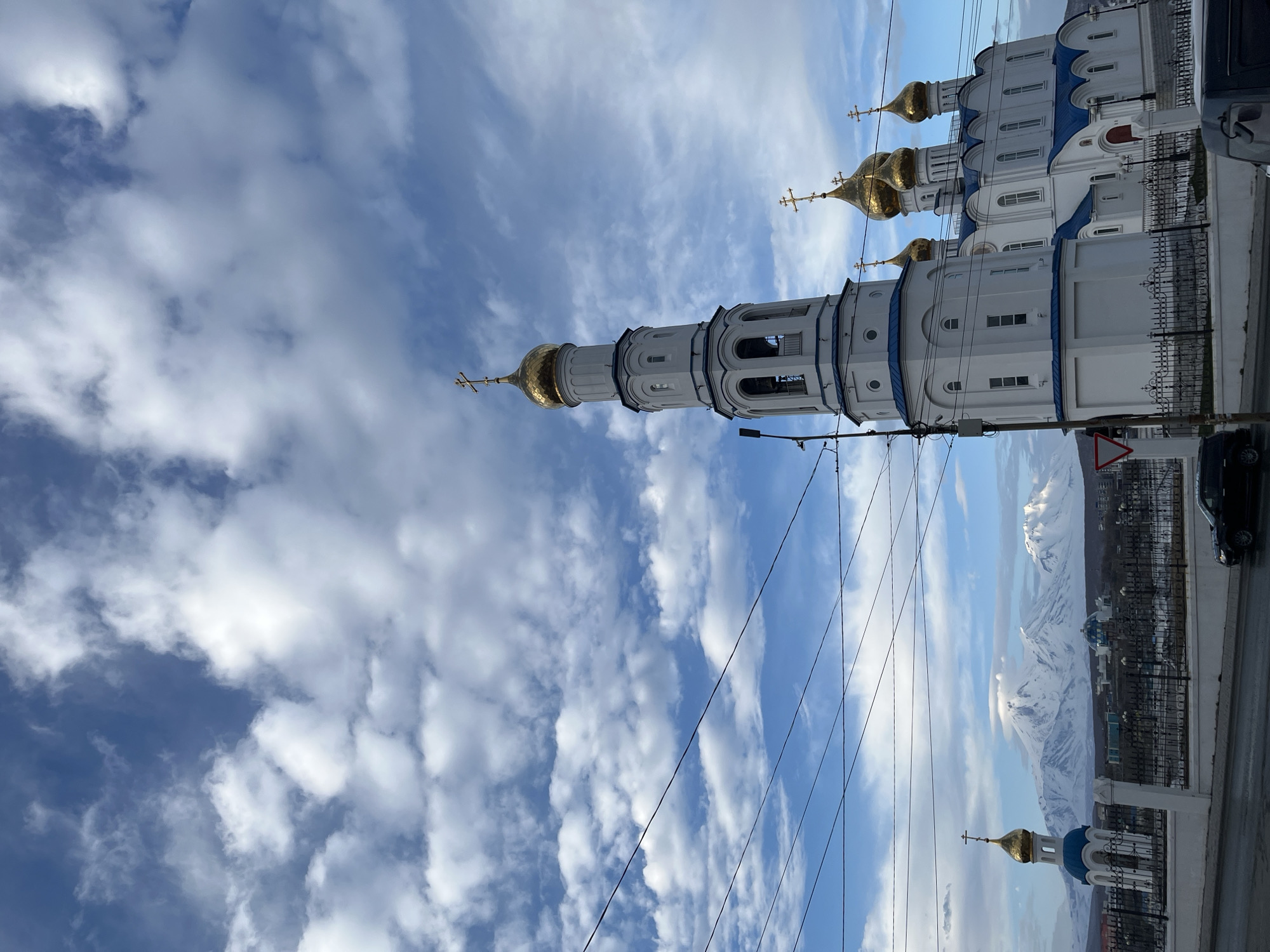 The image shows a church in Petropavlovsk-Kamchatsky, Russia, viewed from a low angle. The church has white walls with blue accents and golden domes topped with Orthodox crosses. The bell tower is prominent. Overhead are power lines. In the background, snow-capped mountains are visible. A black car is partially visible on the right. A street with a metal fence runs along the base of the image. The sky is partly cloudy with patches of blue.