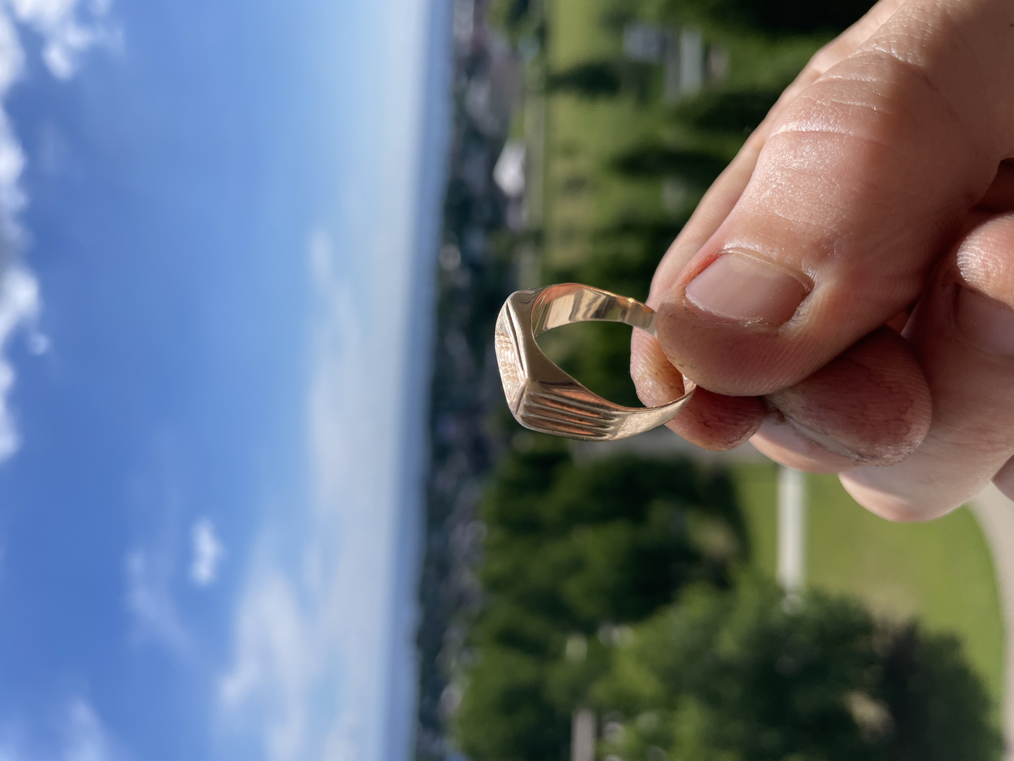 A close-up, eye-level shot shows a person's hand holding a signet ring, with a blurry cityscape in the background under a clear blue sky with scattered white clouds. The signet ring is made of gold and has a rectangular face with vertical lines on the sides. The hand appears to be that of an adult, with visible fingerprints and a clean fingernail. The background shows a landscape with green trees, buildings, and a body of water, suggesting a location on the outskirts of a city or town. The sun appears to be shining brightly, casting highlights on the ring and the hand. The overall impression is one of a casual snapshot taken outdoors on a sunny day.