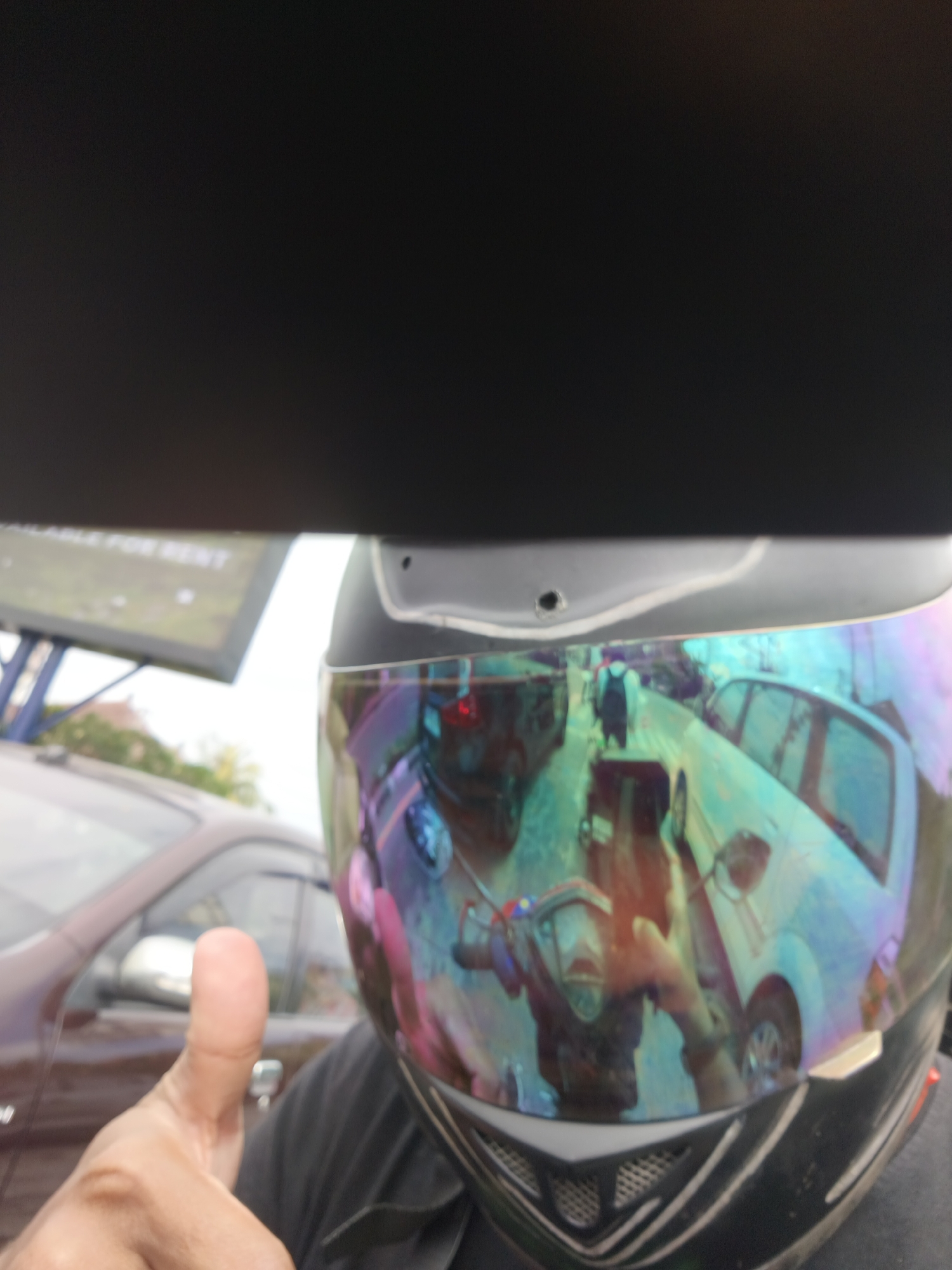 A close-up, over-the-shoulder shot shows a person wearing a black helmet with a reflective, iridescent visor. The visor reflects a street scene in Denpasar, Indonesia. Multiple vehicles, including cars and what appears to be a motorcycle, are visible in the reflection. The person's hand, giving a thumbs-up gesture, is in the foreground, obscuring part of the helmet and visor. A brown car is visible to the left of the frame, and a blurred billboard with text that may say "AVAILABLE FOR RENT" is in the background. The overall image is slightly blurry and the lighting is moderate.