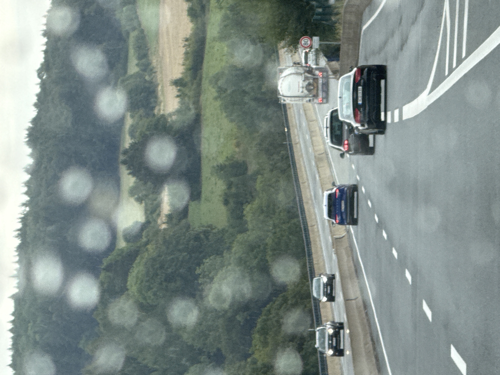 The image shows a view from inside a vehicle, likely taken from a car's windshield, with numerous water droplets obscuring the scene. The view is of a highway with multiple lanes of traffic. In the foreground, there are several cars. Closest to the viewer and in the right lane, a black Audi sedan is visible, with its license plate partially obscured. Further ahead in the center lane, a dark grey car is driving, followed by a blue sedan in the left lane. In the right lane, further ahead, a white tanker truck is visible, with a circular speed limit sign showing "80" next to it. Beyond the highway, the landscape consists of rolling hills covered in dense green trees and fields. The sky appears overcast. The overall scene depicts a highway journey through a rural, wooded area, with the visibility affected by rain or condensation on the viewing surface. This scene is not located in the city of Bad Lauterberg im Harz, Germany; it is a generic highway scene.