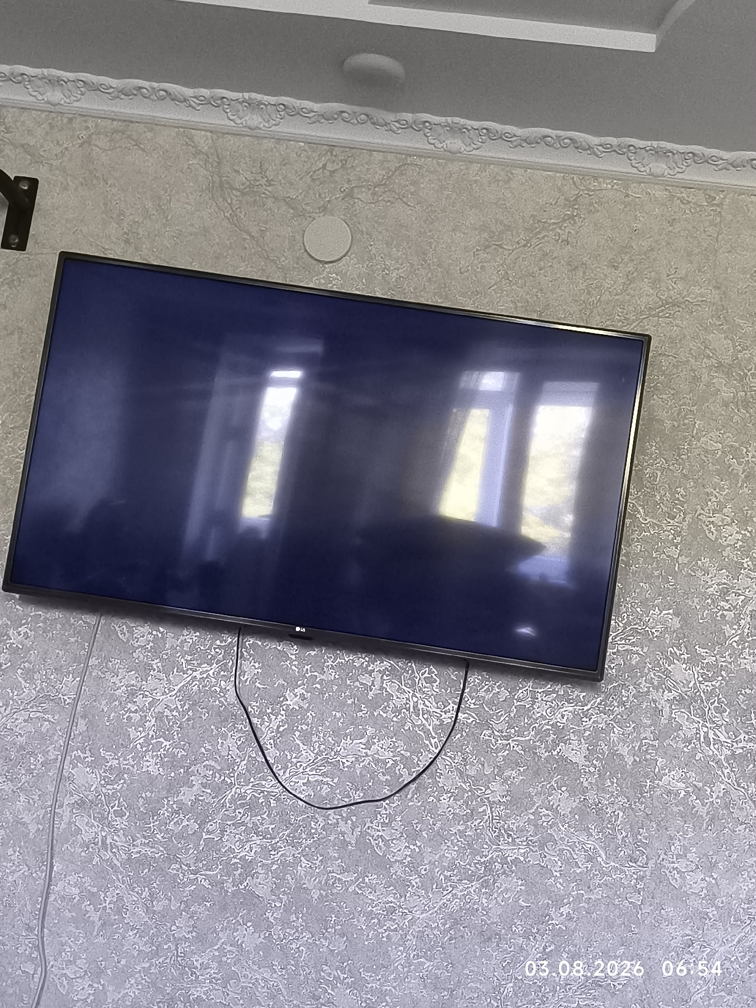 A large, dark television screen is mounted on a textured, light-colored wall, adorned with a decorative molding at the top near the ceiling. The screen reflects the light from a window, creating bright rectangular shapes on its surface. A dark cable is visible hanging from the bottom of the television. In the bottom right corner of the image, a timestamp reads "03.08.2026 06:54". The image appears to be an interior shot of a room. The location in the city of Satbayev, Kazakhstan is not discernible from the visual information provided in the image.