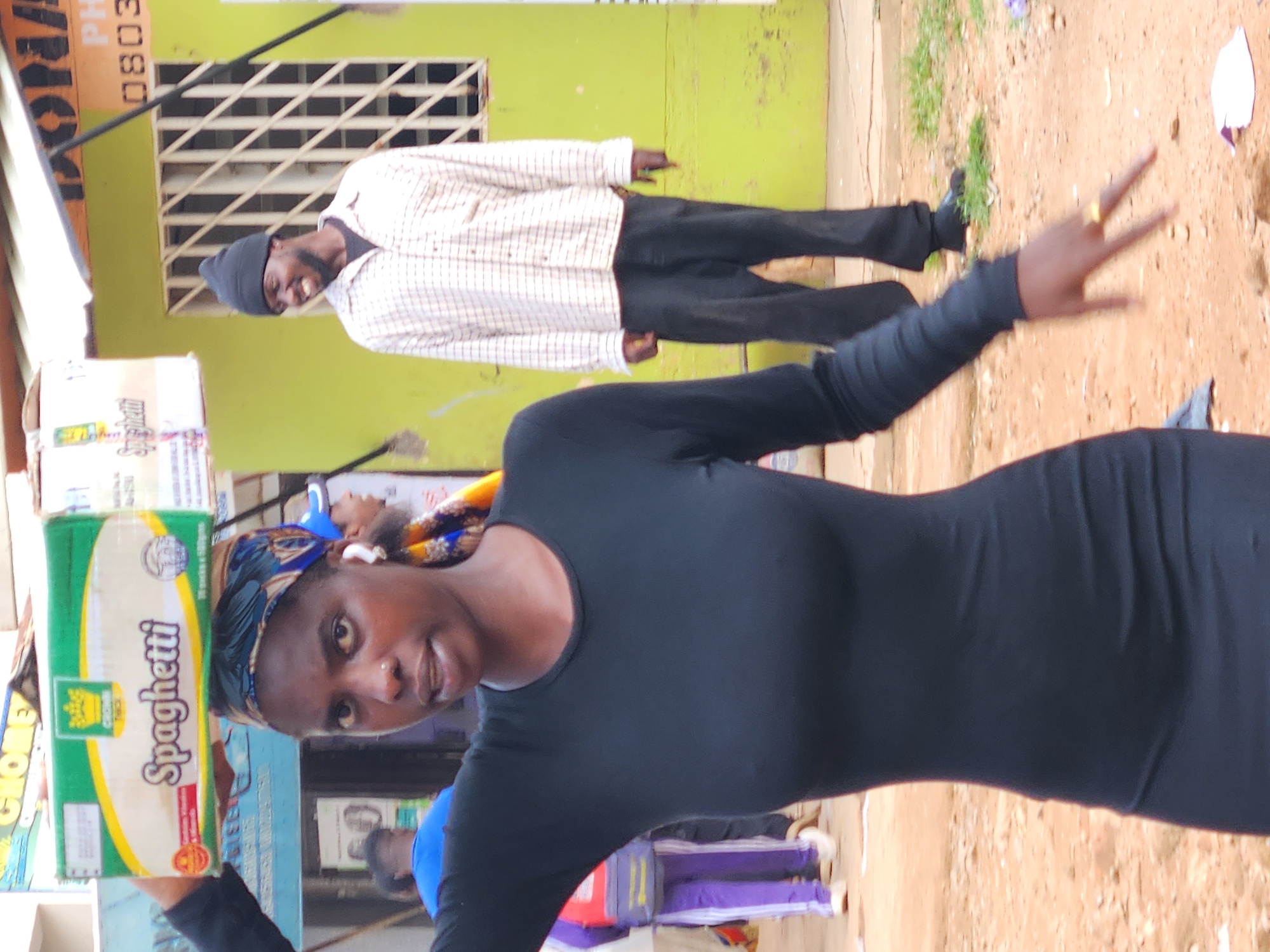 In an outdoor urban environment, a young Black woman stands prominently in the foreground, wearing a fitted black dress and a patterned headscarf. She is carrying a large green and white box of spaghetti balanced on her head. Her left arm is raised, and her hand is visible, adorned with a gold ring. To her right and slightly behind her, a Black man in a plaid shirt and dark pants, wearing a black beanie, smiles. In the background, another person in a blue shirt and a person in a purple tracksuit are partially visible. The ground is dirt, with some patches of dry grass. A light green building with a barred window is visible behind the man, and a street sign with "PH" and numbers is on the building. The overall scene appears to be a street market or a busy thoroughfare.