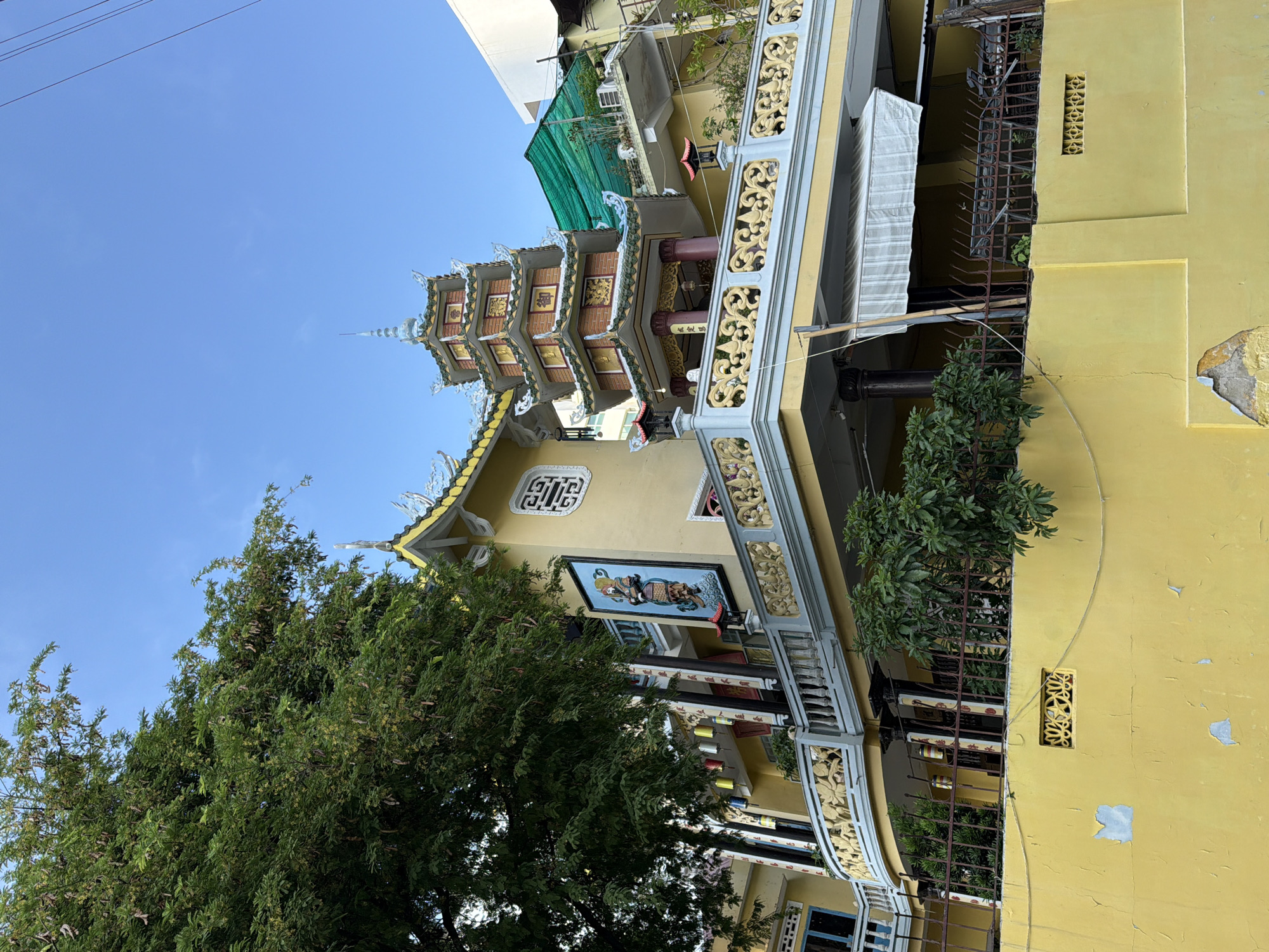 The image depicts a vibrant yellow building with traditional Asian architectural elements, likely a temple or pagoda, situated in Nha Trang, Vietnam. A multi-tiered pagoda with a green tiled roof and ornate golden accents stands prominently on the upper level. The building features decorative balconies with intricate yellow railings, and a large, colorful mural of a figure in traditional attire is displayed on the exterior wall. Lush green trees partially obscure the building, and a clear blue sky serves as the backdrop. The lower section of the building is a bright yellow wall with some peeling paint, and a decorative ventilation grille. The scene suggests a sunny day in a densely populated urban environment, common in Vietnamese cities.