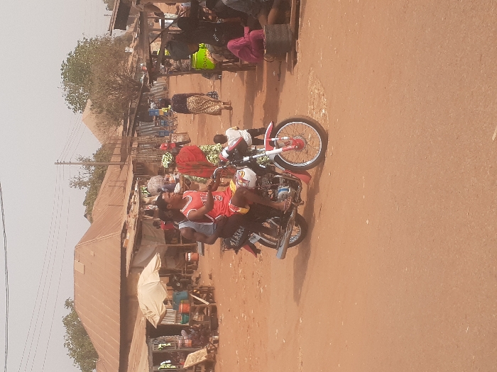 The image portrays an outdoor street or market scene in Gwadanye, Nigeria, under a hazy sky. A reddish-brown dirt road dominates the foreground. Two adult males are riding a white and red motorcycle, observed from the front-right. The driver wears a red tank top with a white bull logo and red shorts, looking slightly right. The passenger, seated behind, wears a light-colored tank top and dark shorts. The motorcycle features a "LAGIRI" sticker on its rear.
To the right of the motorcycle, several individuals are present, including a woman in a red headscarf and patterned dress, another woman in a leopard-print skirt walking away from the viewer, and a small child in a white shirt standing near the road. A bright green gas cylinder is visible next to an informal market stall. To the left, multiple makeshift market stalls with various goods are set up, one shaded by a large, light-colored umbrella.
In the background, numerous buildings with weathered, corrugated metal roofs and rough walls are visible. Utility poles and overhead wires crisscross the upper portion of the frame, with sparse-leaved trees interspersed among the structures.