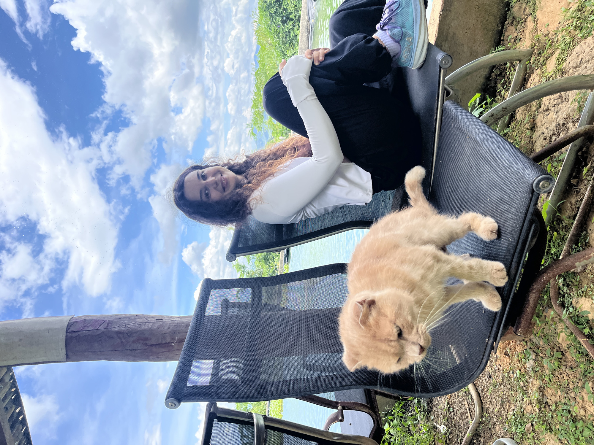 A young woman with curly brown hair is seated on a black outdoor chair. She is wearing a white long-sleeved shirt and black pants. Her left arm is resting on her knee, and she is looking at the camera with a smile. In front of her, on another black outdoor chair, is a light orange cat. The cat is standing and looking down. The chairs are situated outdoors next to a body of water, possibly a pool or a lake, with lush green vegetation and distant mountains visible under a partly cloudy sky. The location is specified as Sơn Điền, Vietnam. The scene appears to be a relaxed, outdoor setting.