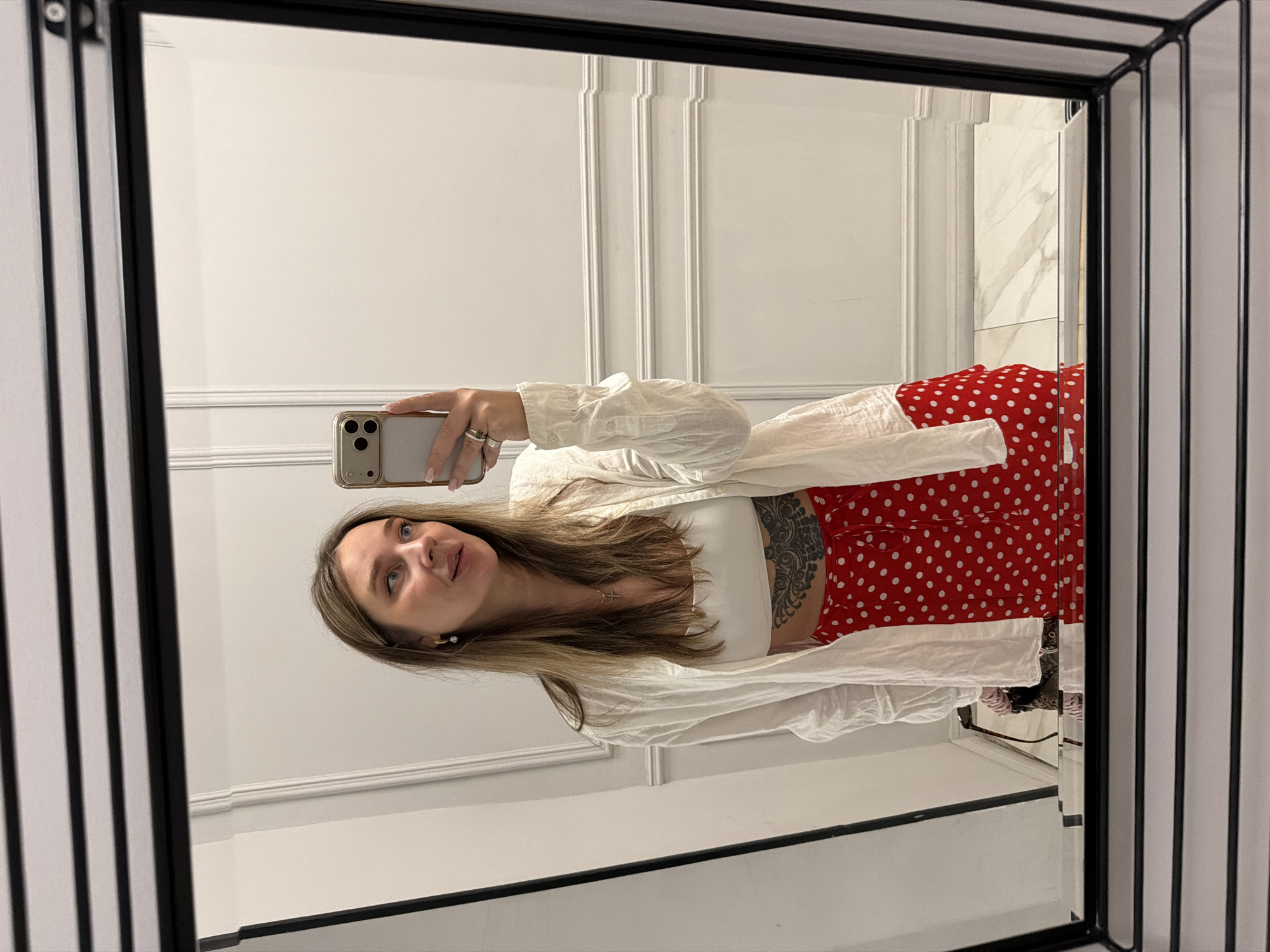 A woman is taking a mirror selfie in what appears to be a dressing room or bathroom. The woman is wearing a white crop top, a red and white polka-dot skirt, and a white, unbuttoned, long-sleeved shirt. The phone in her hand is an iPhone, and a tattoo is visible on her left side. The room has white walls with molding details and marble-tiled floors. The mirror has a black multi-lined frame. There is no evidence to suggest the location is in Moscow, Russia.