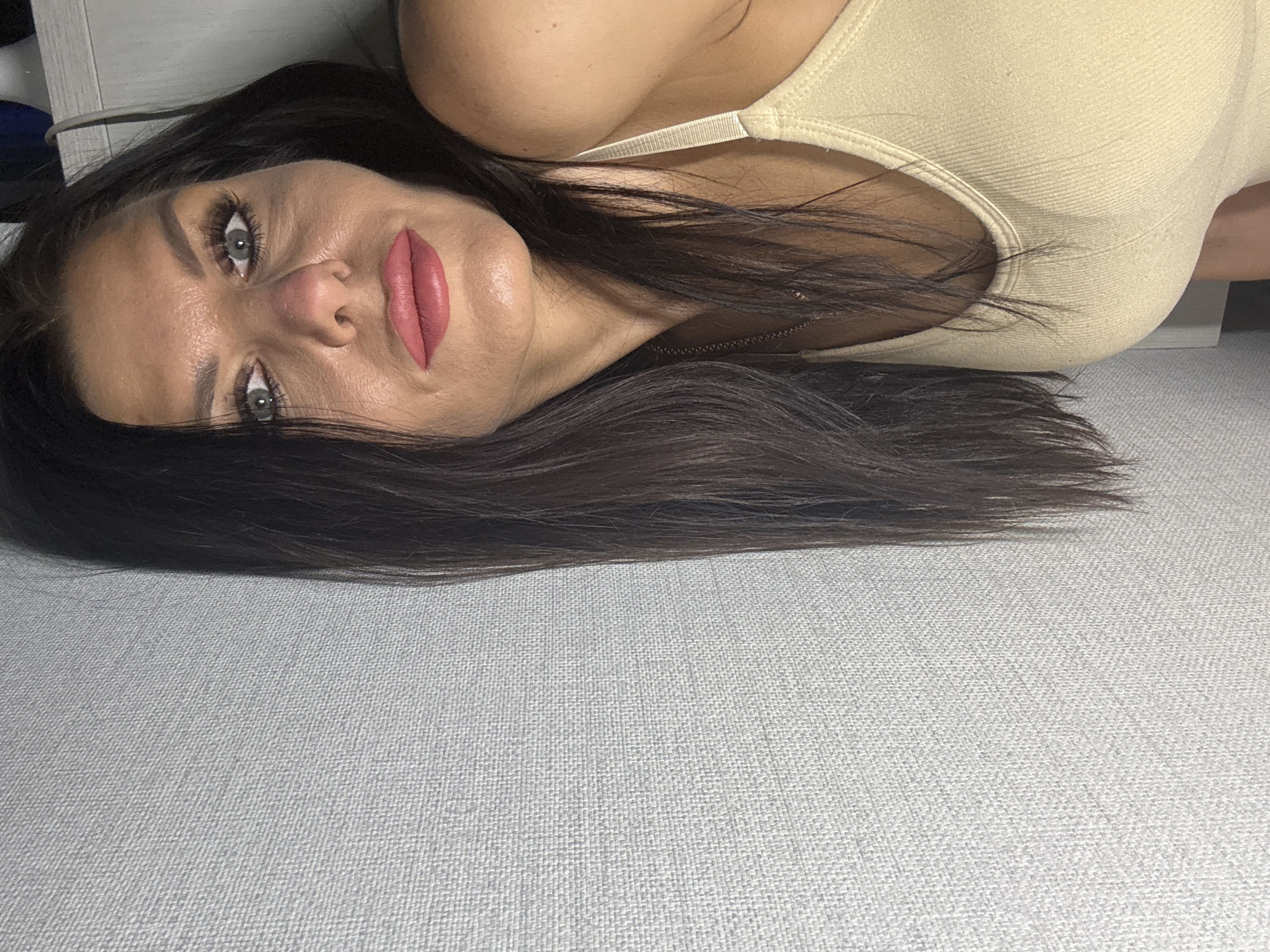 The image is a close-up portrait of a woman lying horizontally across the frame, looking directly towards the viewer. She has long, dark hair that spreads across a light grey, subtly textured fabric surface, likely a sofa or bed. Her features are prominent, with full, bright pink matte lipstick. Her eyes are light-colored, possibly blue or grey, framed by dark, well-defined eyebrows and noticeable mascara or false eyelashes. A delicate, thin metallic necklace is visible around her neck. She wears a light beige or cream-colored ribbed top or bralette, with part of her shoulder and upper chest visible. The lighting is soft and even, creating a clear view of her skin texture and makeup. Her expression is neutral and direct, suggesting a posed shot. The overall impression is one of a relaxed, intimate setting. Although not depicted in the image itself, the provided context specifies the location as Chaikovsky, Russia. There is no visible text in the image.